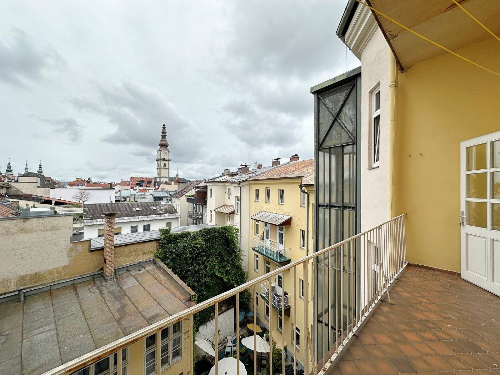 Balkon mit Blick auf die Dächer der Stadt und einen Kirchturm unter bewölktem Himmel.
