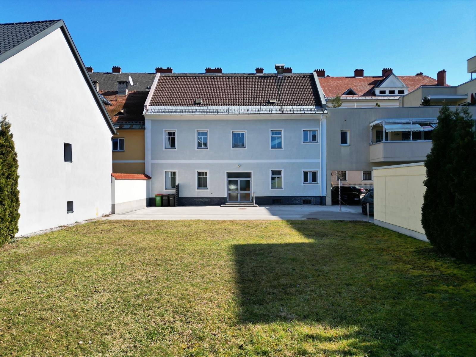 Rückansicht des Gebäudes mit einer großen Rasenfläche und angrenzenden Gebäuden unter blauem Himmel.