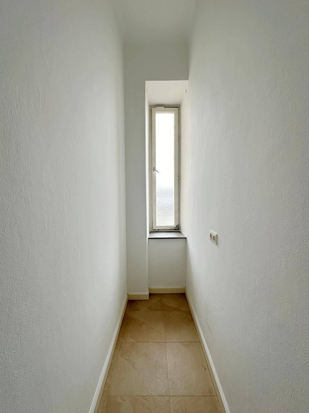 Schmaler Flur mit hellem Fliesenboden und einem hohen Fenster, das Tageslicht spendet.