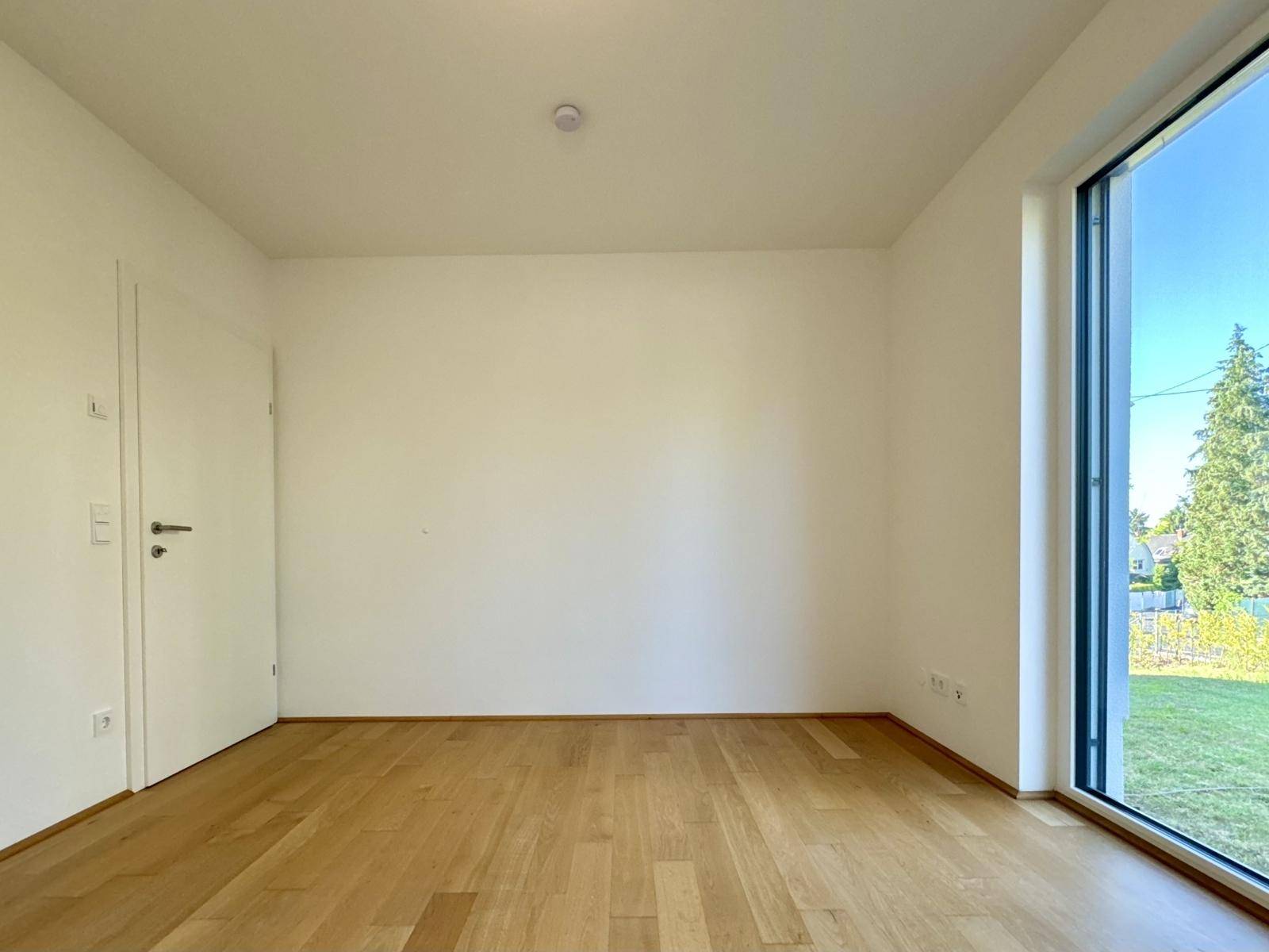 Helles Zimmer mit Holzboden und bodentiefem Fenster, das viel Tageslicht hereinlässt.
