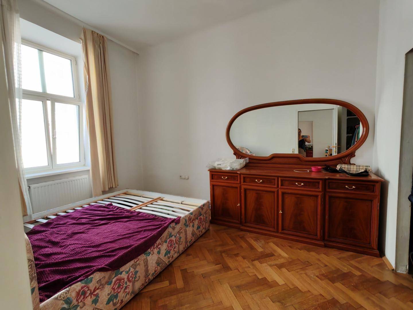 Schlafzimmer mit Fischgrätparkett, großem Fenster und klassischer Holzkommode mit ovalem Spiegel.