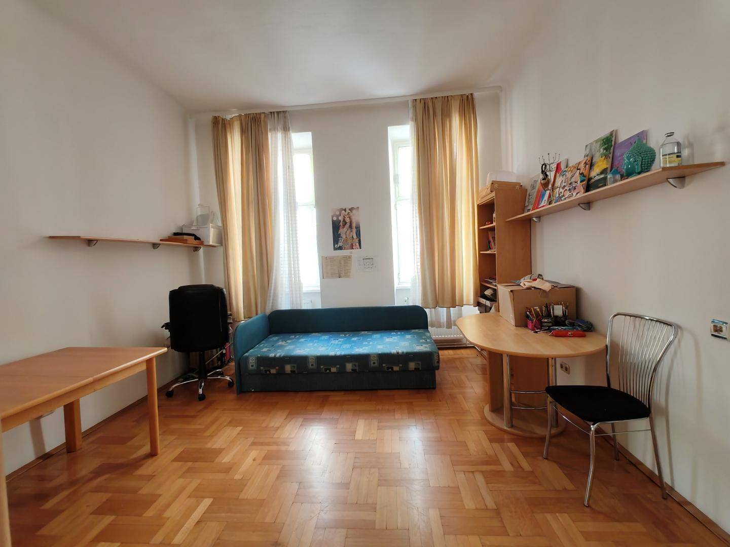 Helles Zimmer mit Fischgrätparkett, Schlafsofa und einem Bereich, der als Büro genutzt werden kann.