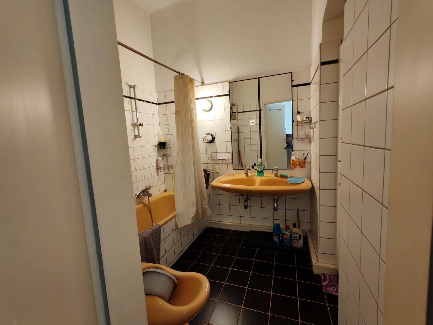 Badezimmer im 80er-Jahre-Stil mit gelber Badewanne, Waschbecken und schwarzen Bodenfliesen.