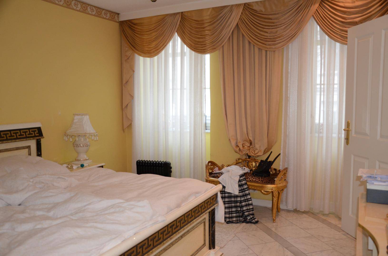 Schlafzimmer mit gelben Wänden, großem Bett und eleganten Vorhängen vor den Fenstern, mit Fliesenboden.