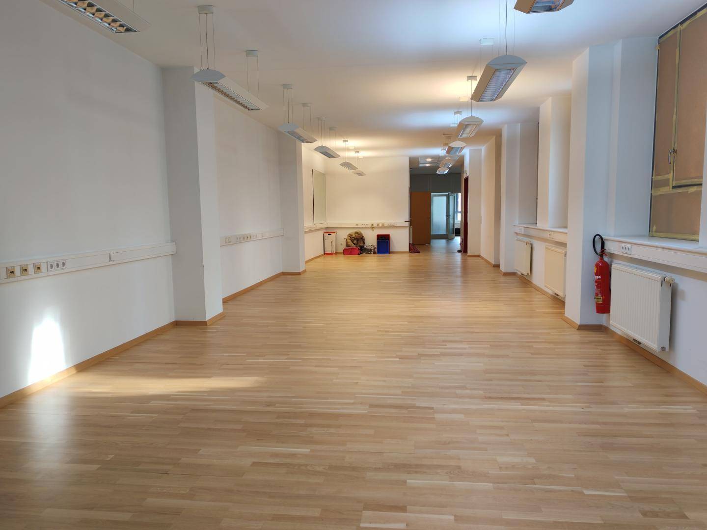 Geräumiger, heller Raum mit Holzfußboden und vielen Steckdosen, ausgestattet mit Heizkörpern und einem Feuerlöscher.