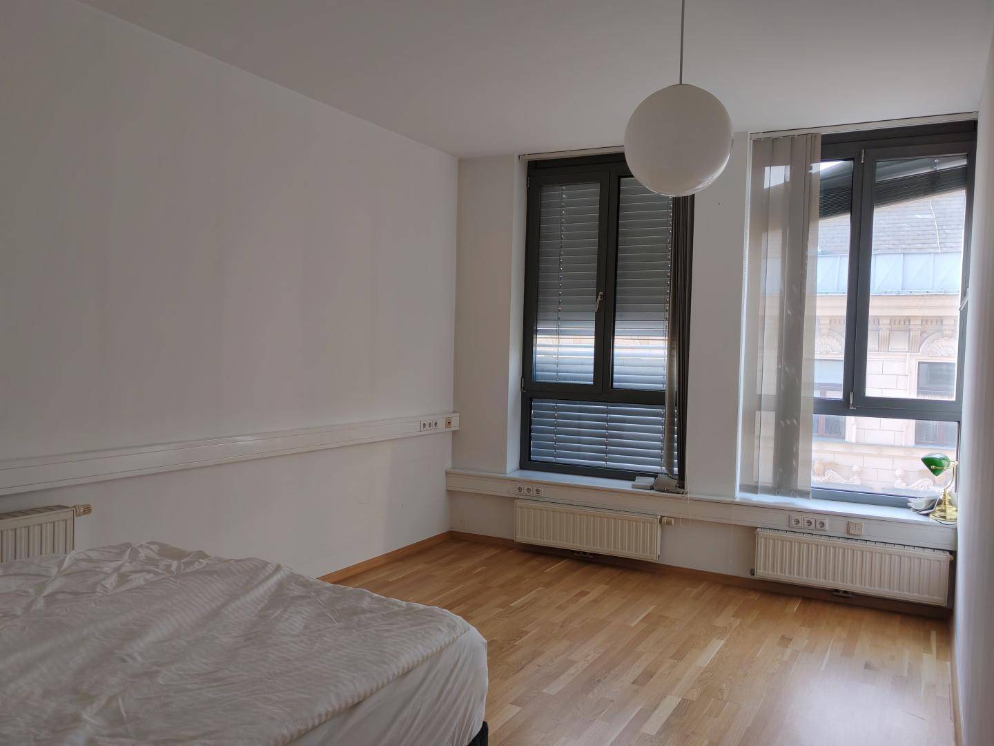 Geräumiges Schlafzimmer mit großen Fenstern, hellem Holzfußboden und Heizkörpern unter den Fensterbänken.