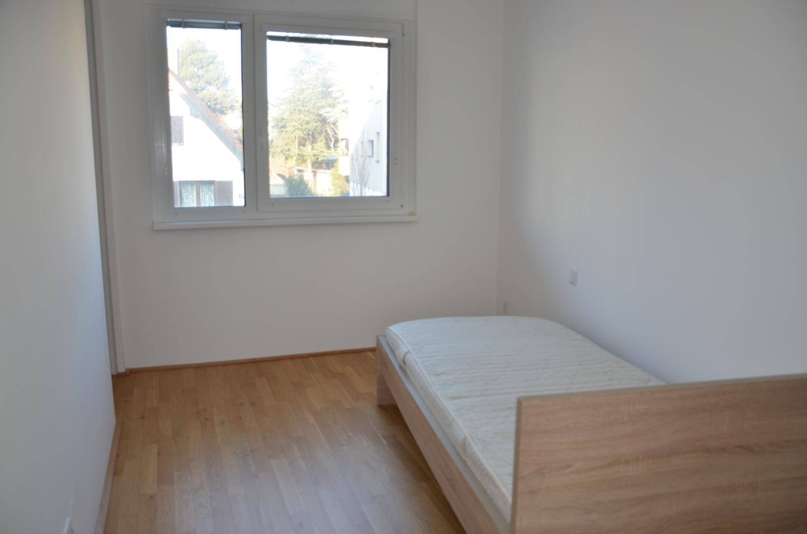 Schlafzimmer mit großem Fenster, Holzparkettboden und viel natürlichem Lichteinfall.