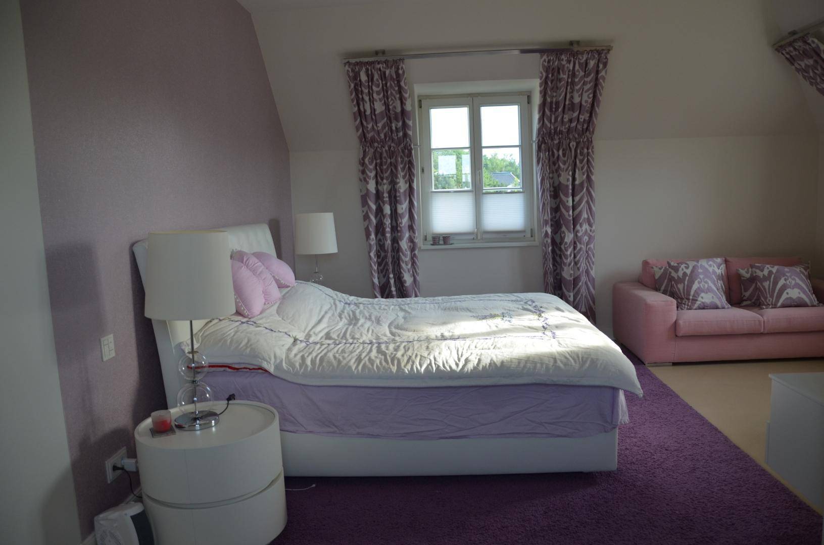 Gemütliches Schlafzimmer in Lila-Tönen mit Doppelbett, Sofa und Teppichboden.