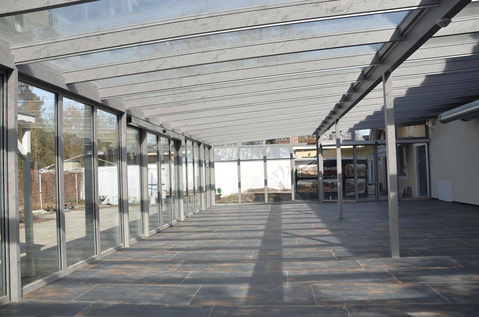 Langer, verglaster Anbau mit Metallrahmen und dunklem Fliesenboden, ideal als Wintergarten oder überdachter Gang.