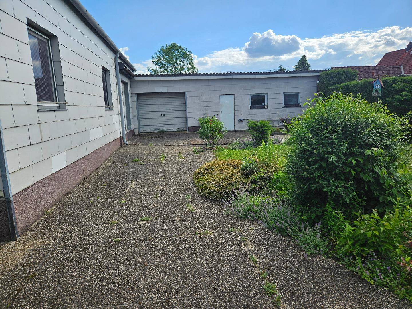Gepflasterter Bereich neben dem Haus mit angrenzendem Garten und einer Garage im Hintergrund.