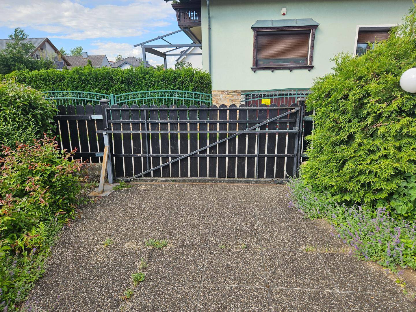 Einfahrtstor aus Metall und Holz vor einem Haus mit grüner Hecke und Rollläden.