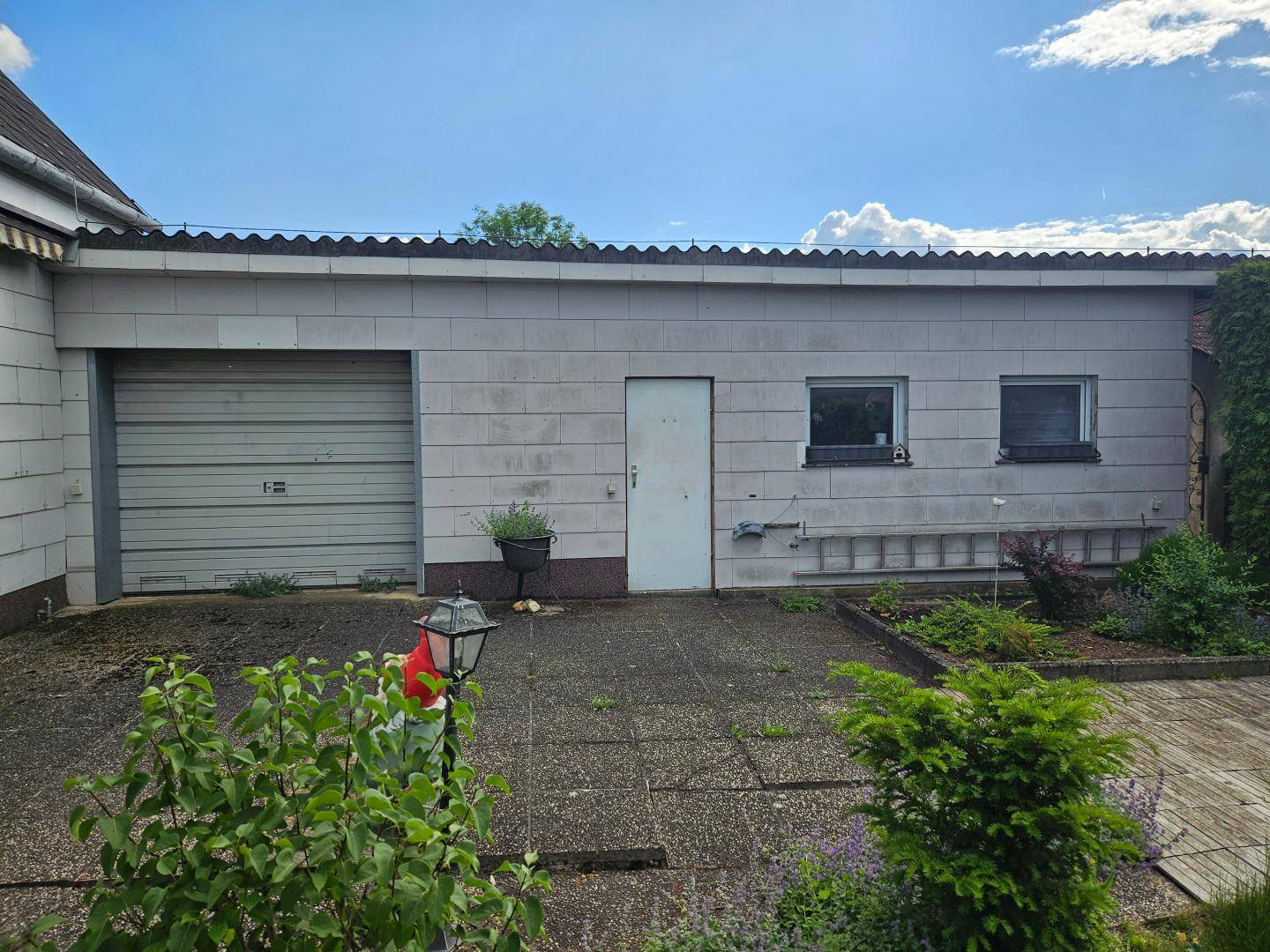 Ansicht der Garage mit Rolltor und zwei Fenstern, umgeben von einem gepflasterten Hof.