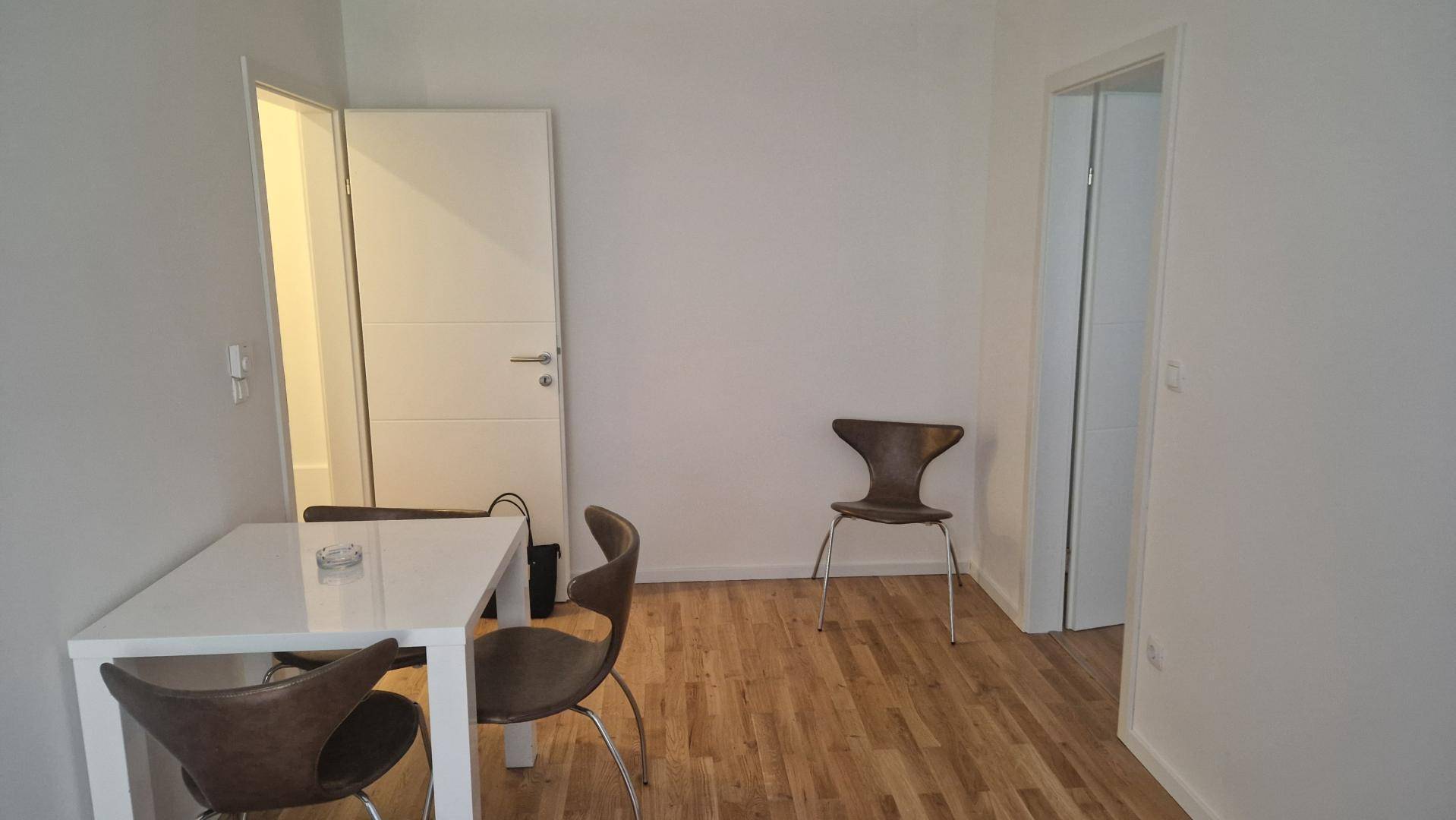 Heller Raum mit Holzboden, einem weißen Tisch und zwei Stühlen, sowie zwei Türen.