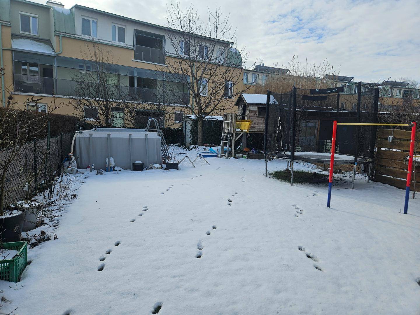 Verschneiter Garten mit Aufstellpool, Trampolin und Spielhaus hinter dem Gebäude.