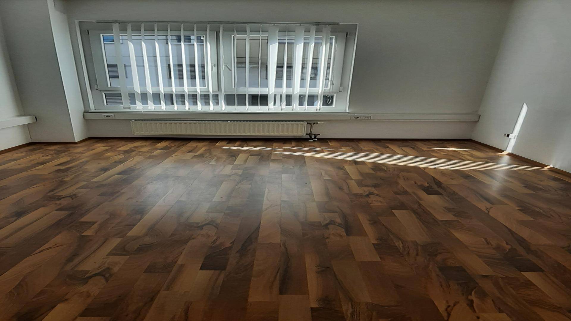Heller Büroraum mit schrägem Dach und einem großen Fenster mit Jalousien, das viel Licht spendet.