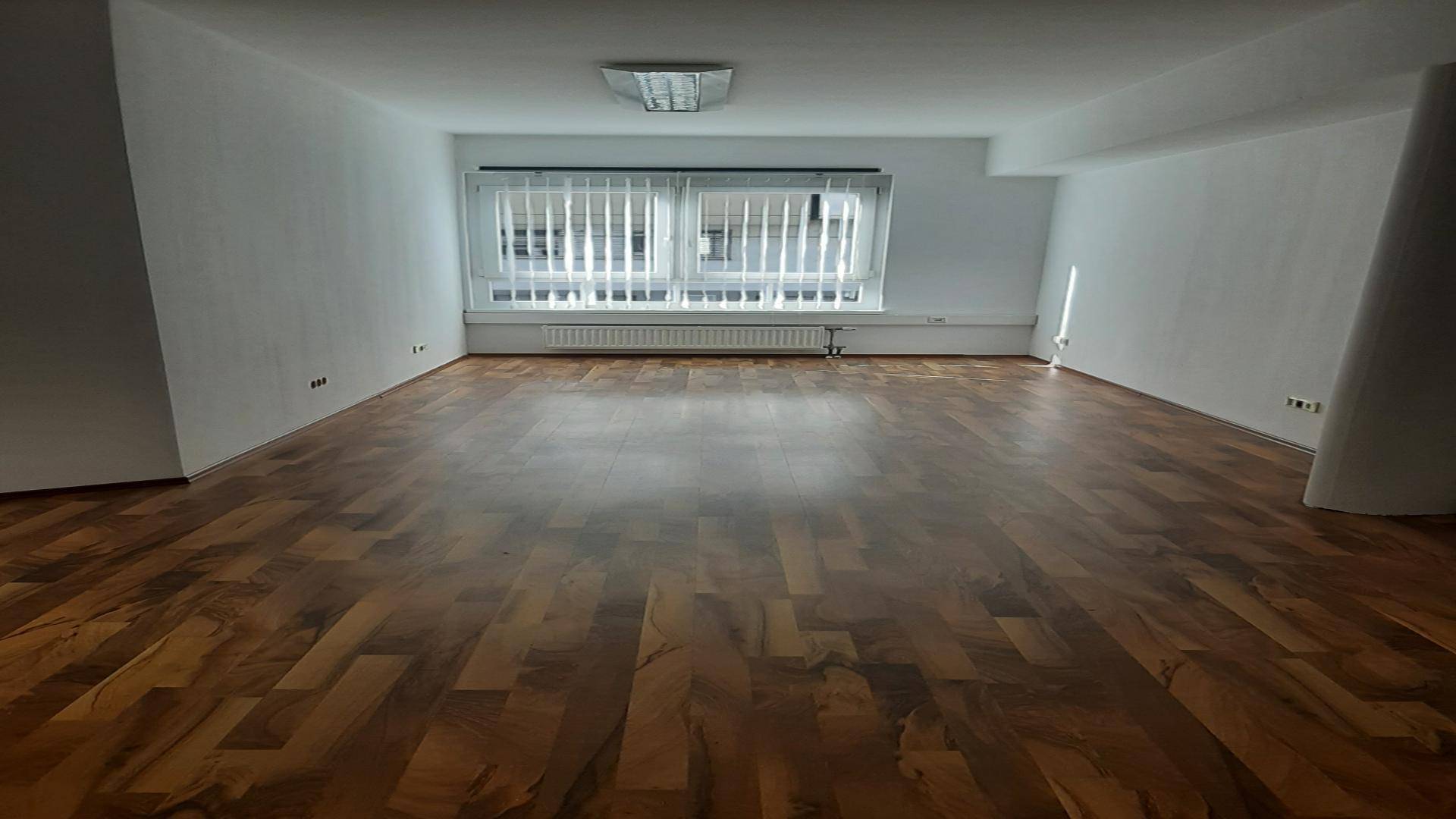 Langer Büroraum mit Holzparkett und einem großen Fenster mit Jalousien, das den Raum erhellt.