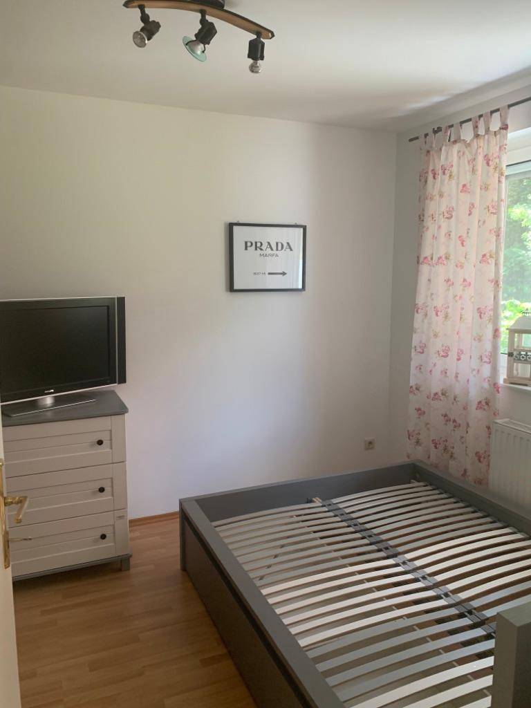 Schlafzimmer mit einem Bettgestell, Kommode und Fenster mit floralen Vorhängen.