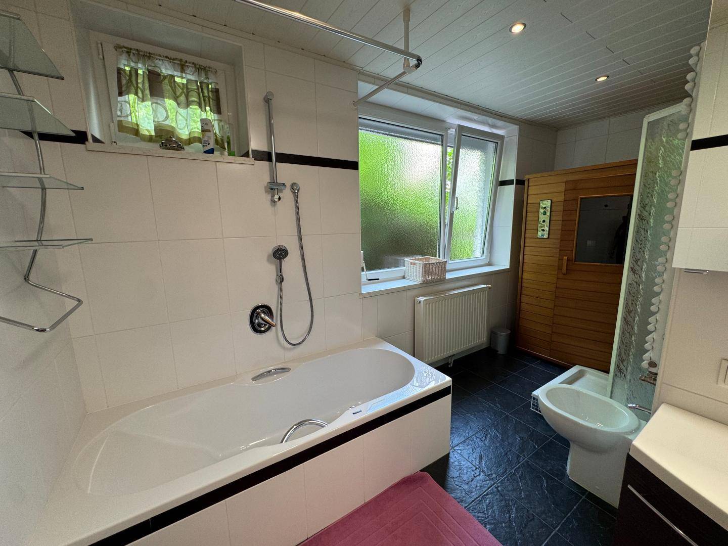 Geräumiges Badezimmer mit Badewanne, Fenster und einer sichtbaren Sauna im Hintergrund.