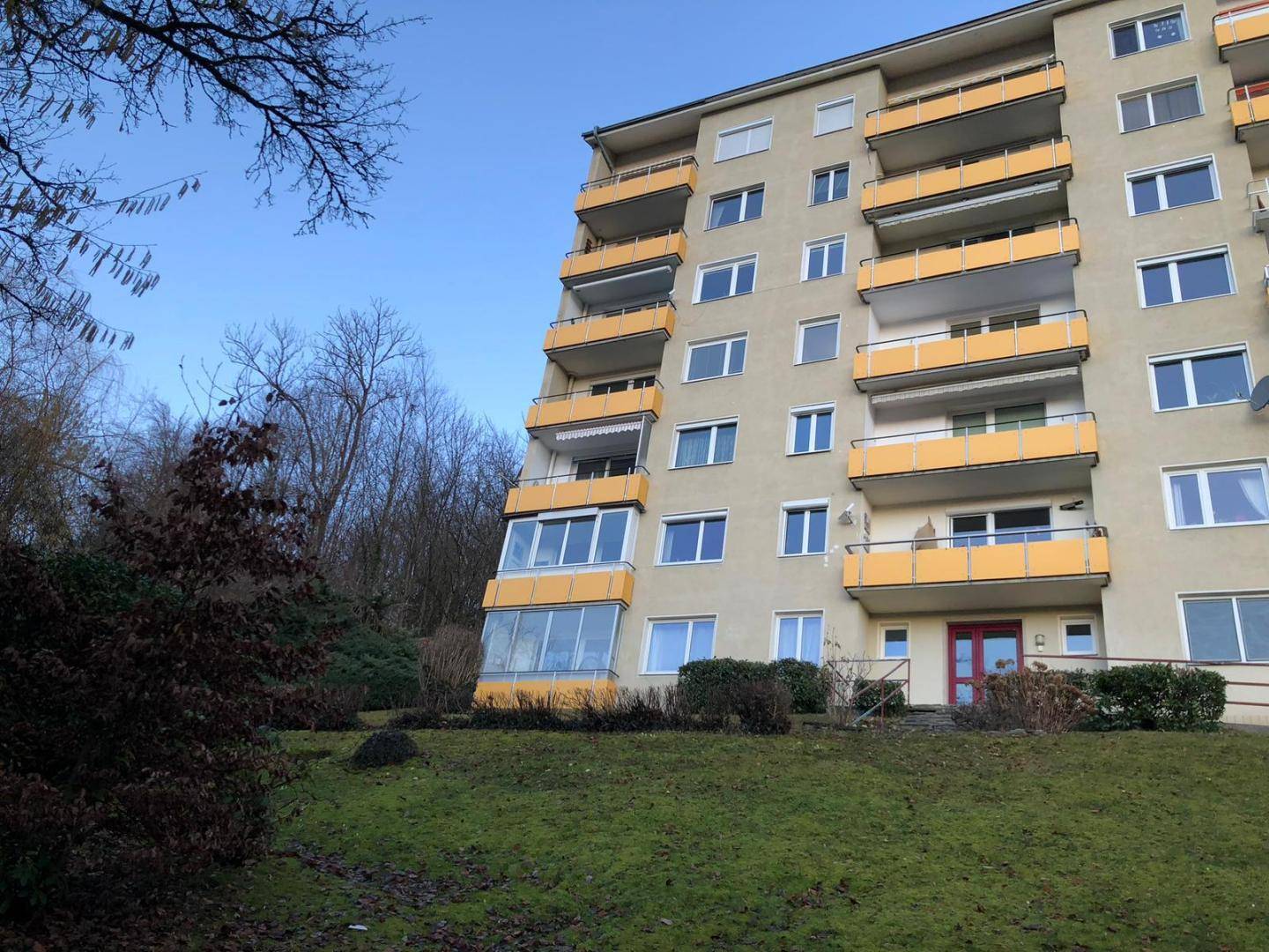 Mehrstöckiges Apartmentgebäude mit gelben Balkonen, eingebettet in eine grüne Hanglandschaft mit Bäumen.