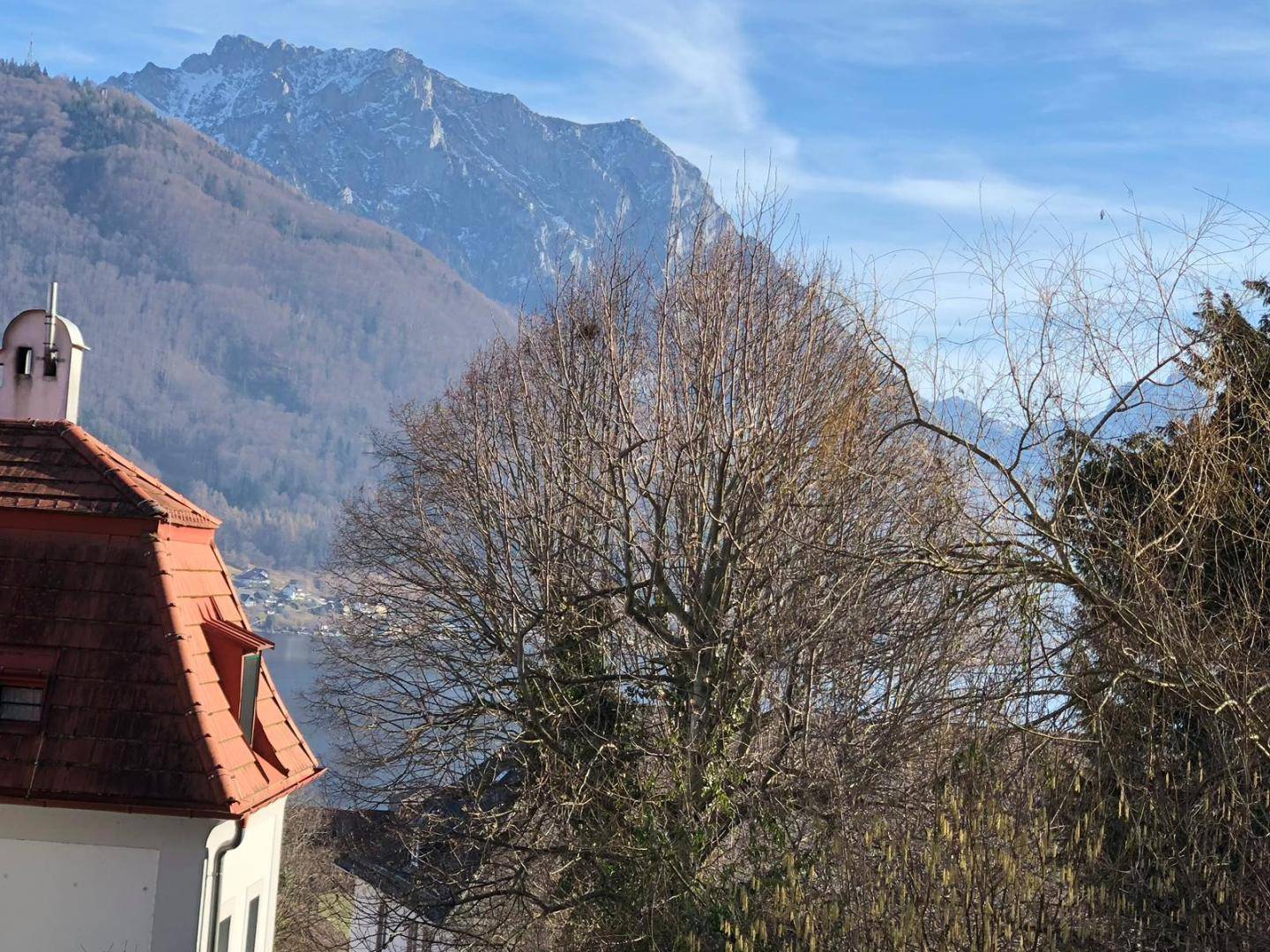 Panoramablick auf einen See und schneebedeckte Berge, umrahmt von einem historischen Gebäude und Bäumen.
