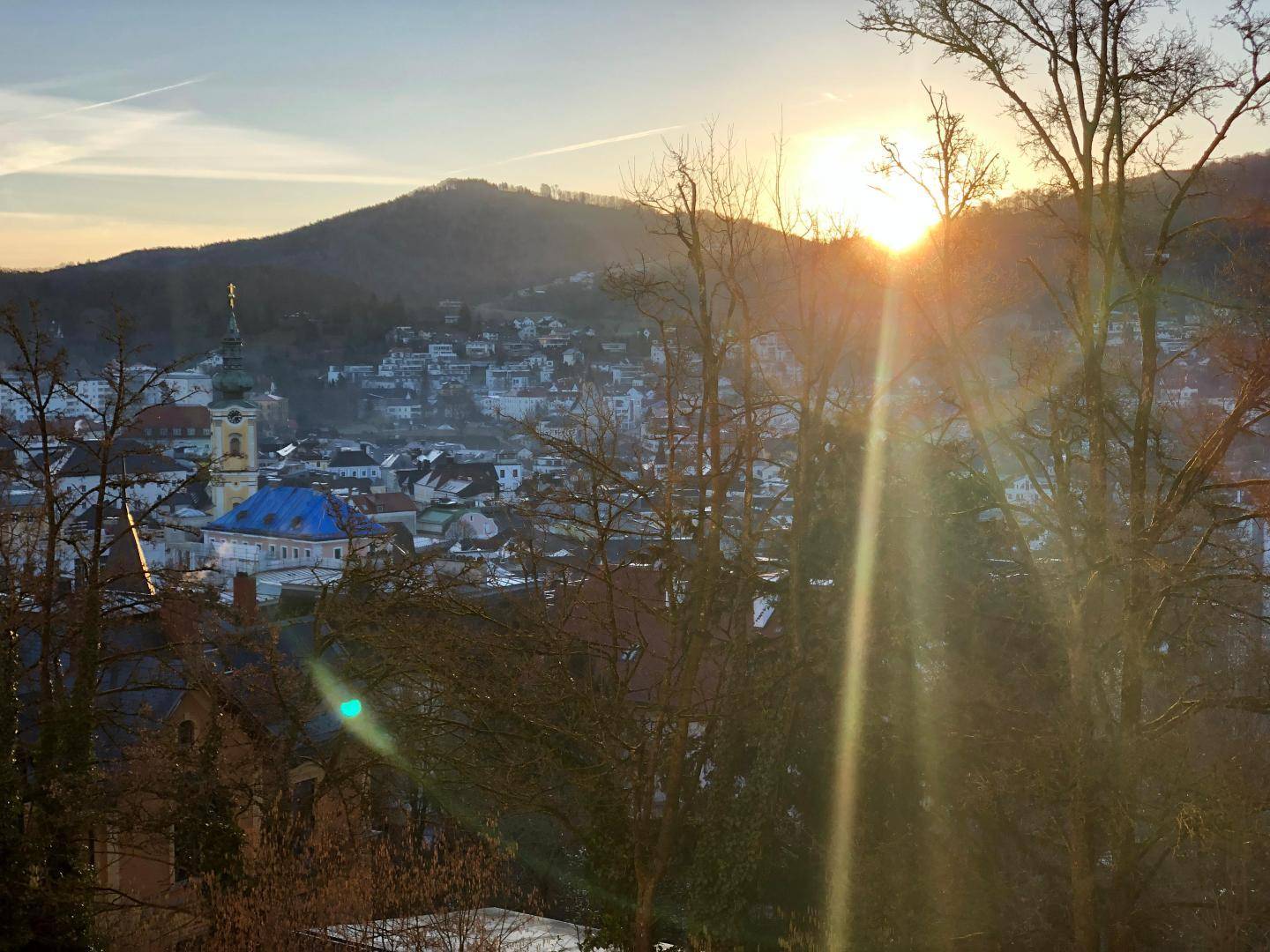Sonnenuntergang über einer malerischen Stadt mit Hügeln im Hintergrund, durch kahle Bäume gesehen.