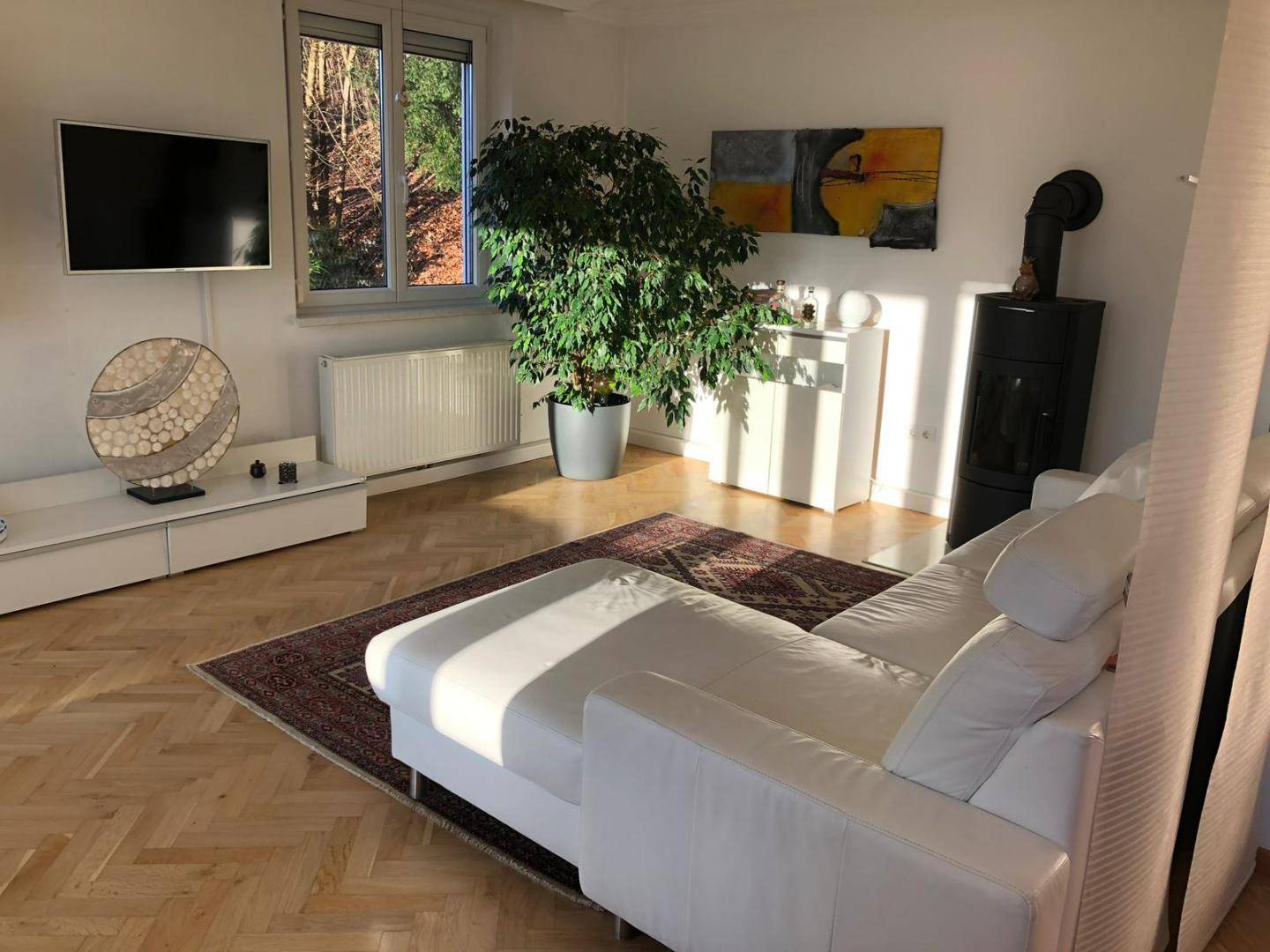 Gemütliches Wohnzimmer mit Kaminofen, Fernseher und großem Fenster, das viel Tageslicht hereinlässt.