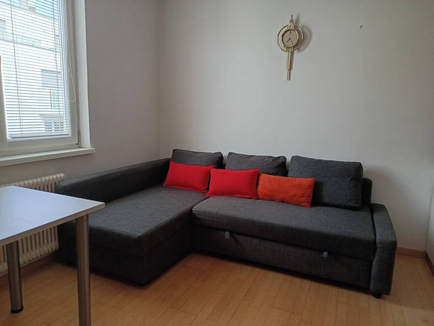 Gemütliches Wohnzimmer mit grauem Ecksofa, hellem Holzfußboden und Fenster mit Jalousien.