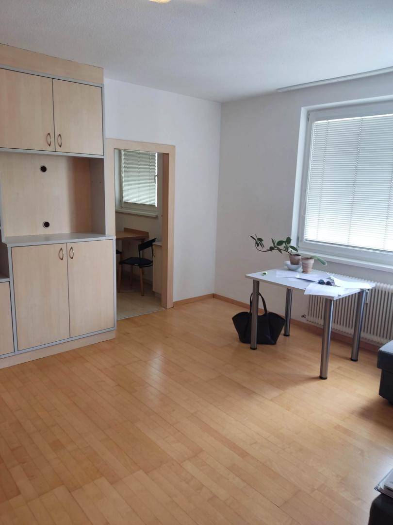 Heller Wohnbereich mit Holzfußboden, großem Fenster und Zugang zu einem weiteren Zimmer.
