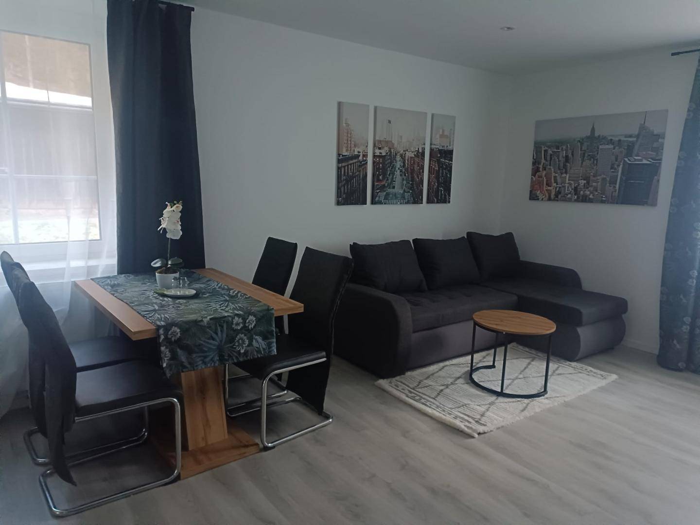 Gemütlicher Wohn- und Essbereich mit dunklem Sofa, Esstisch und Stadtbild-Wanddekoration.