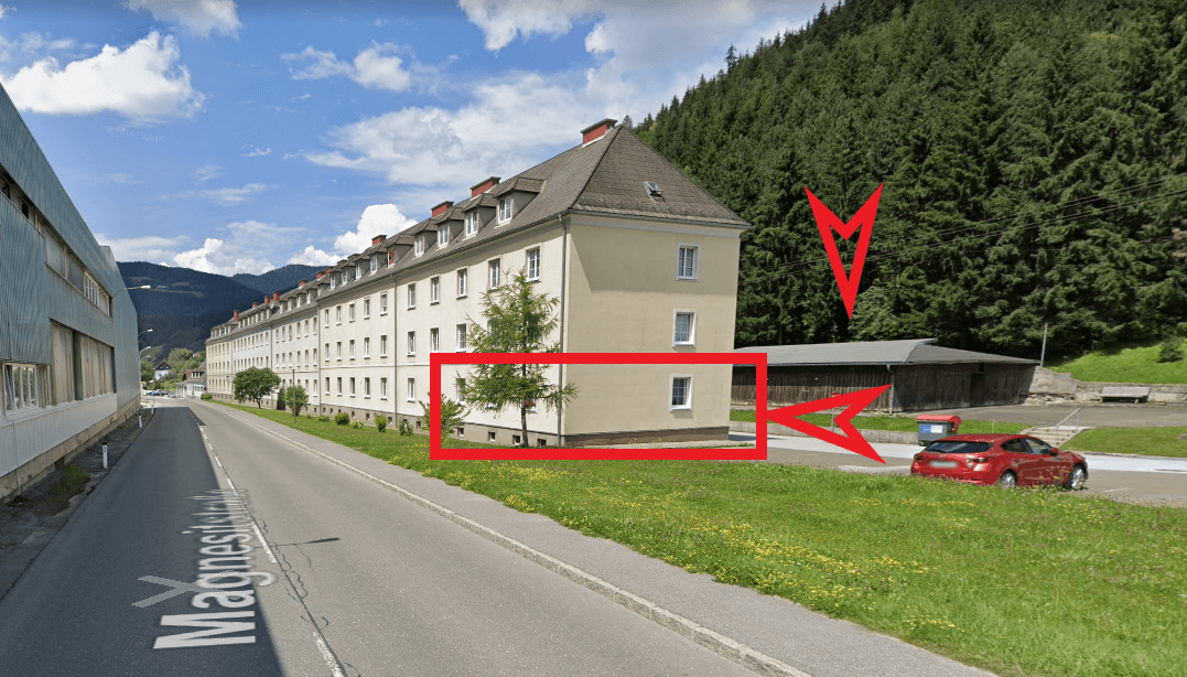 Mehrfamilienhaus an einer Straße mit bewaldetem Hügel im Hintergrund, markiert mit roten Pfeilen.