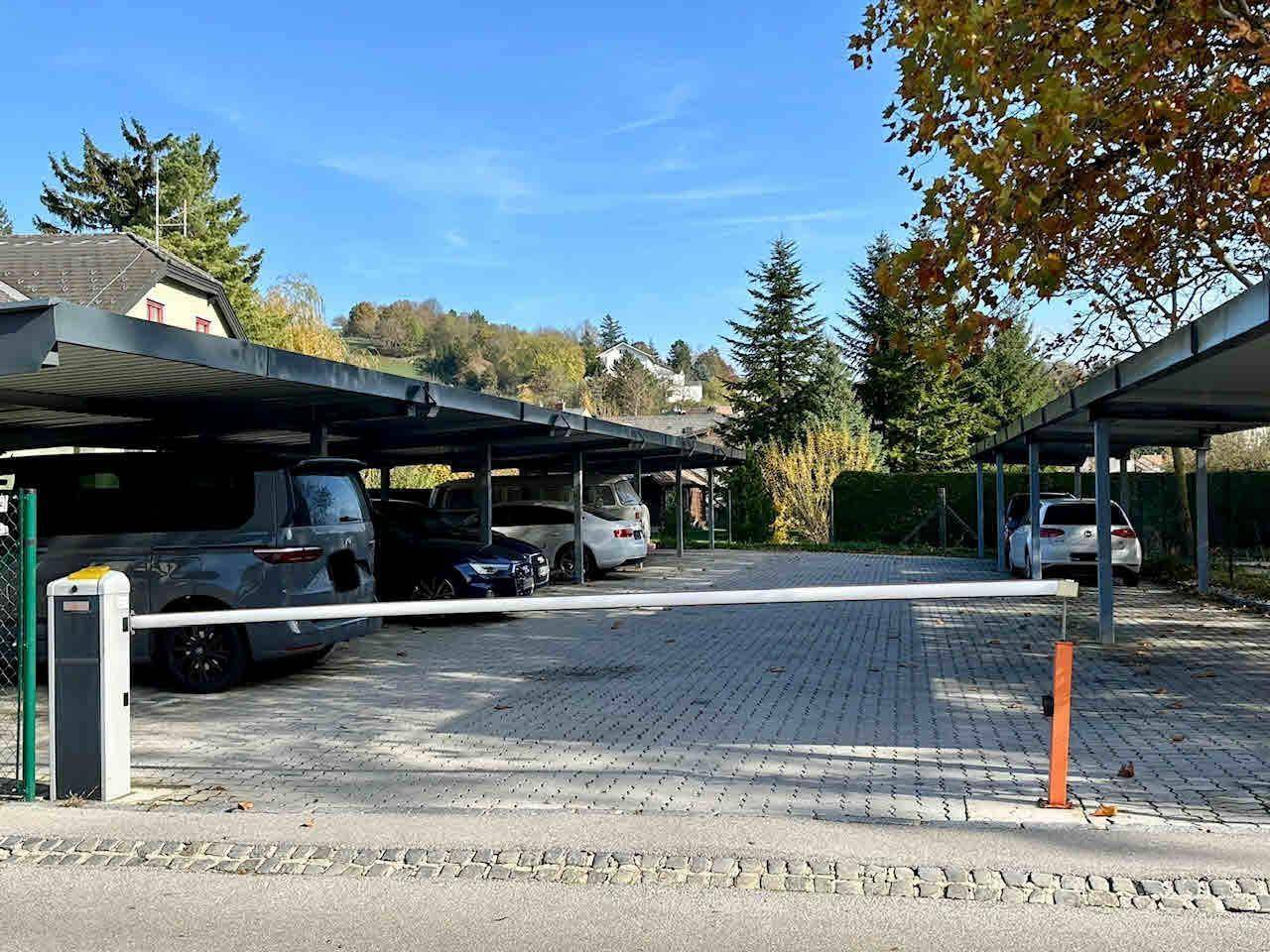 Überdachte Parkplätze mit mehreren Fahrzeugen und geschlossener Schranke, umgeben von Bäumen.
