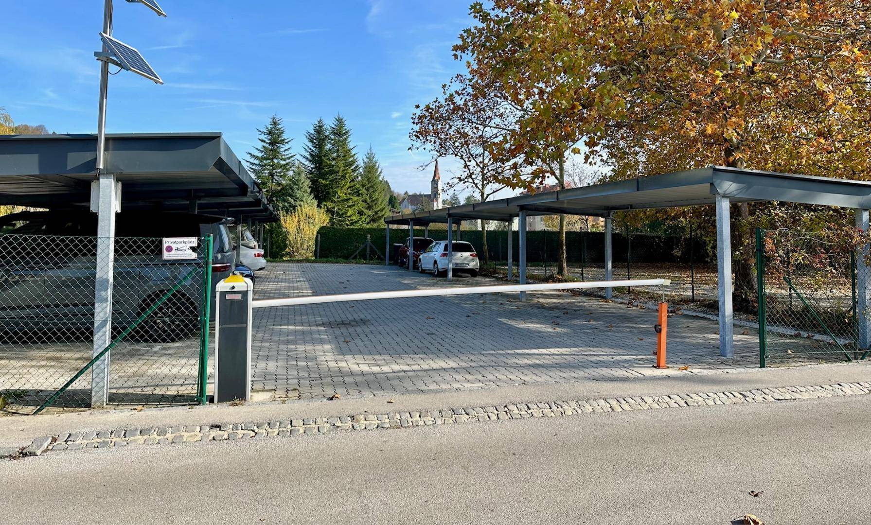 Eingangsbereich des Parkplatzes mit Schranke und überdachten Stellplätzen unter herbstlichen Bäumen.