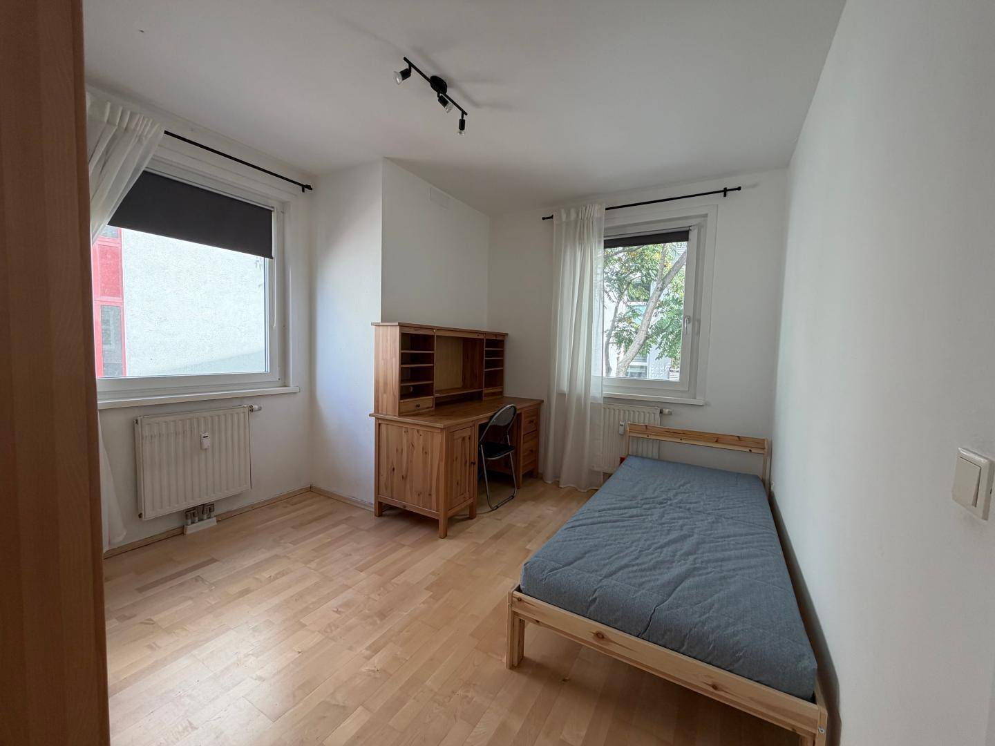 Helles Zimmer mit Einzelbett, Holzschreibtisch und zwei Fenstern.