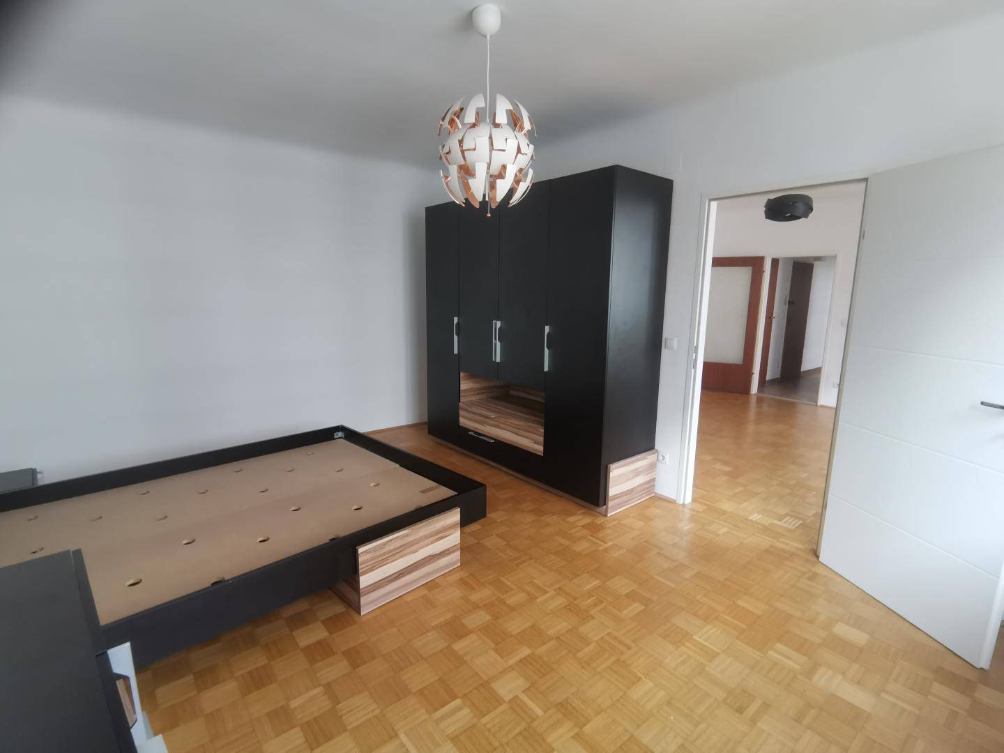 Schlafzimmer mit Parkettboden, dunklem Kleiderschrank und moderner Deckenleuchte.