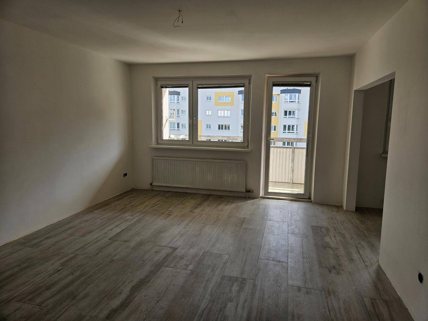Geräumiges Wohnzimmer mit hellem Fliesenboden, großem Fenster und Zugang zu einem Balkon.