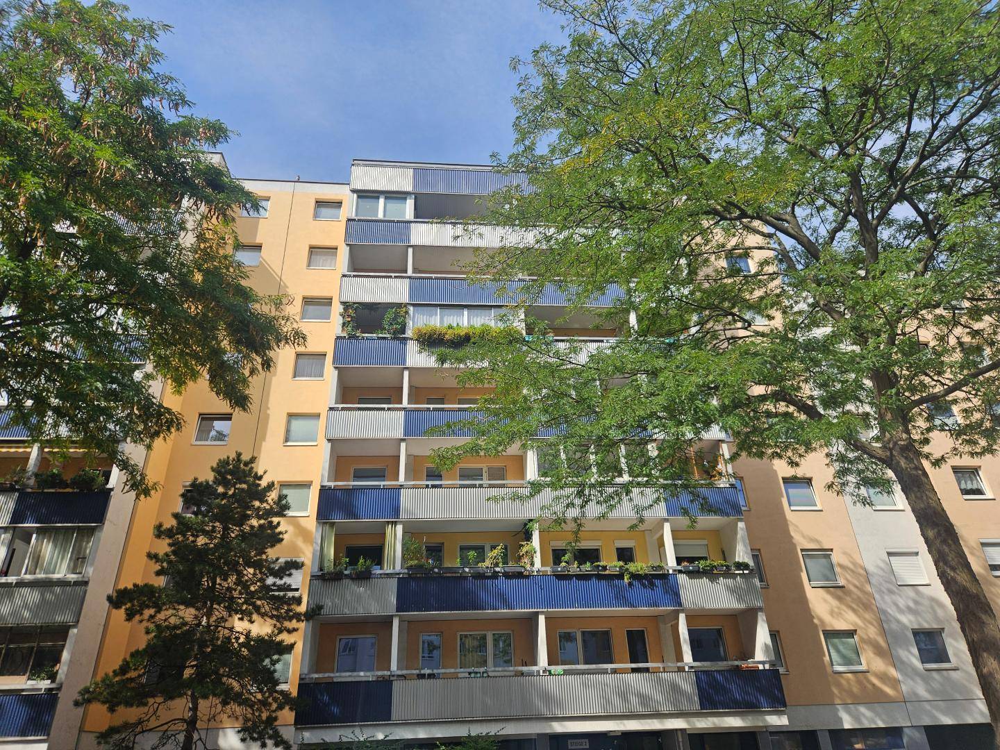 Ansicht eines Mehrfamilienhauses mit zahlreichen Balkonen und grünen Bäumen im Vordergrund.