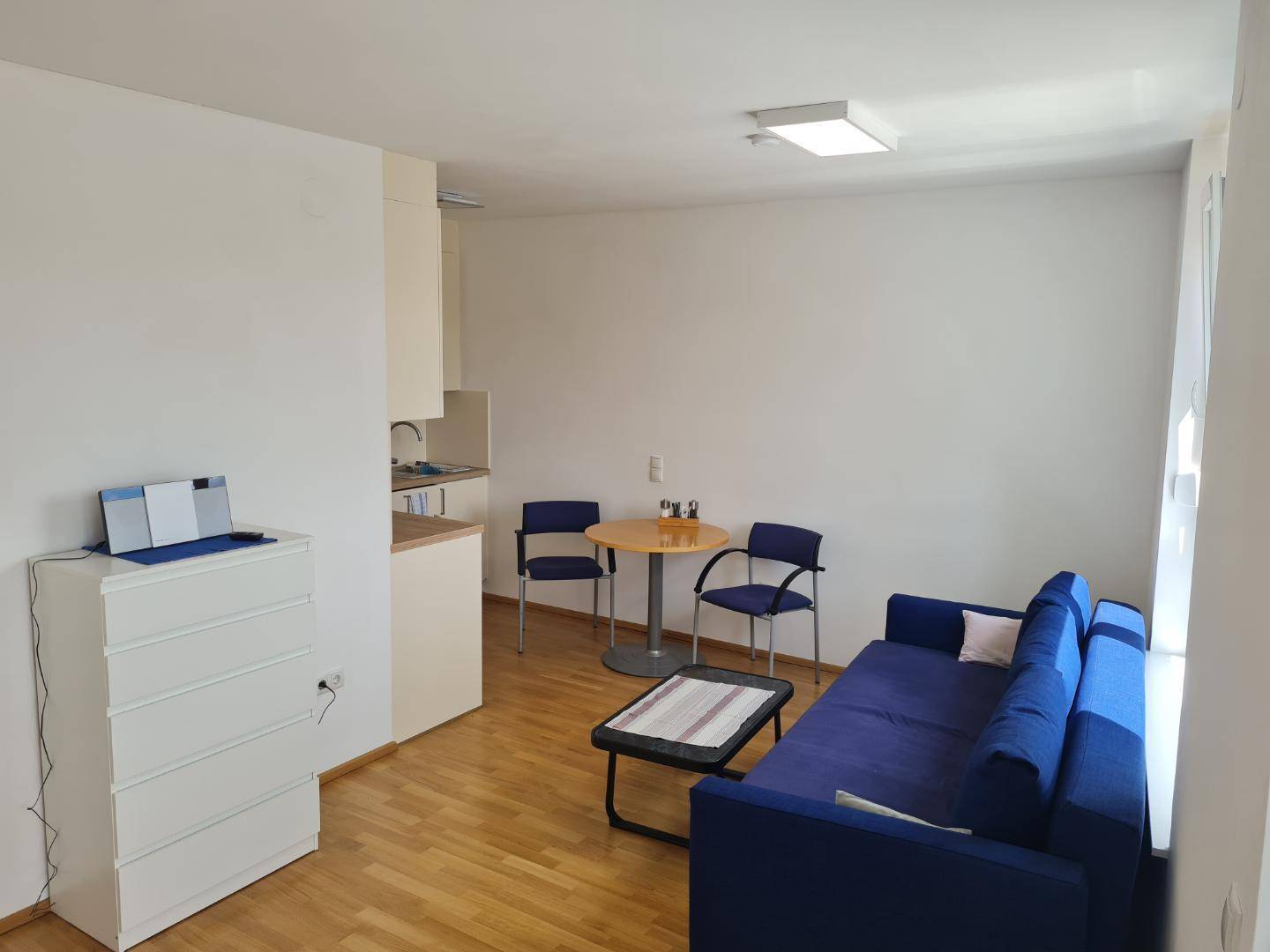 Heller Wohnbereich mit blauem Sofa, Esstisch und integrierter Küchenzeile.