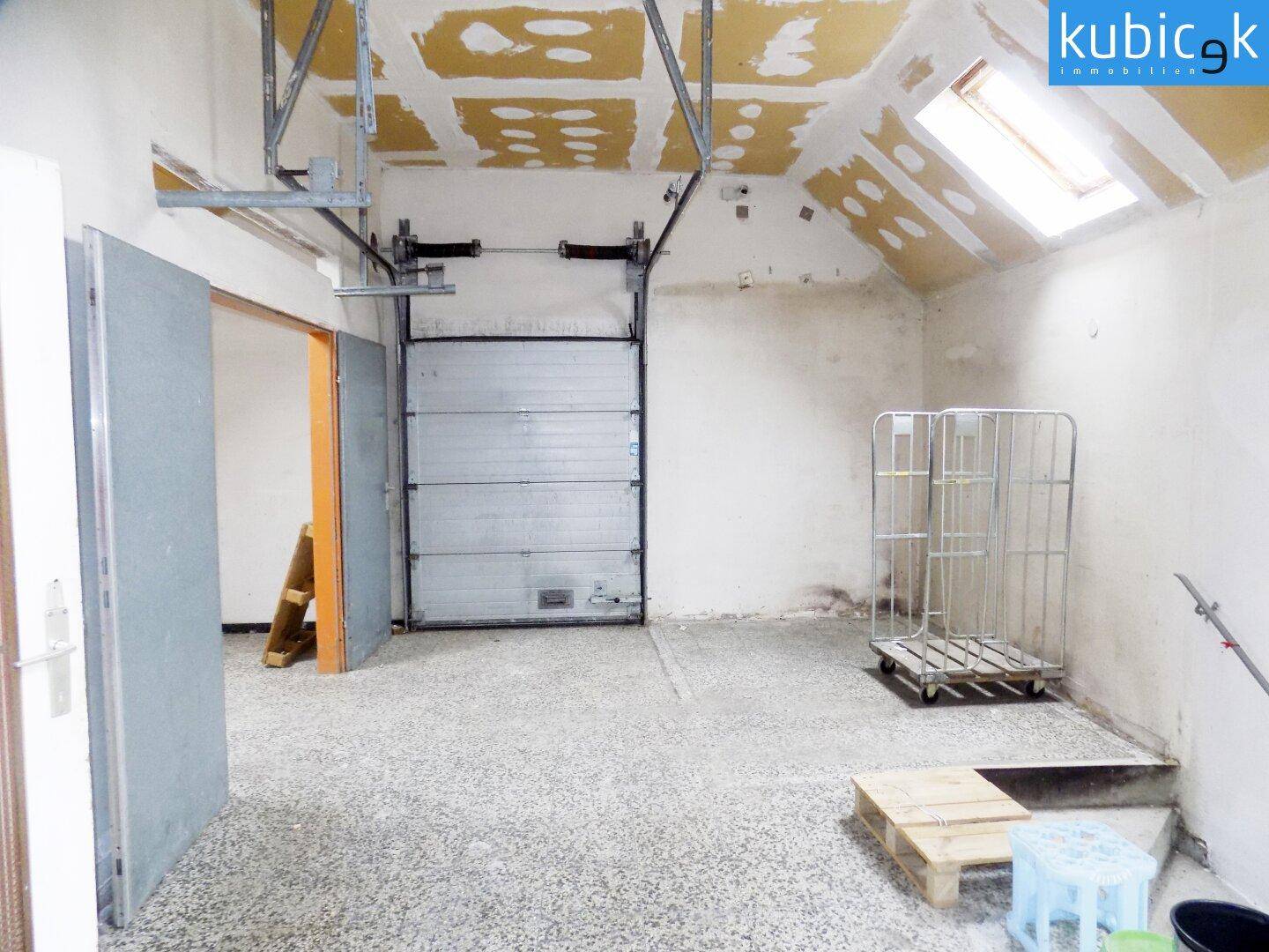 Geräumige Garage oder Lagerhalle mit Rolltor, schräger Decke, Dachfenster und gesprenkeltem Boden.