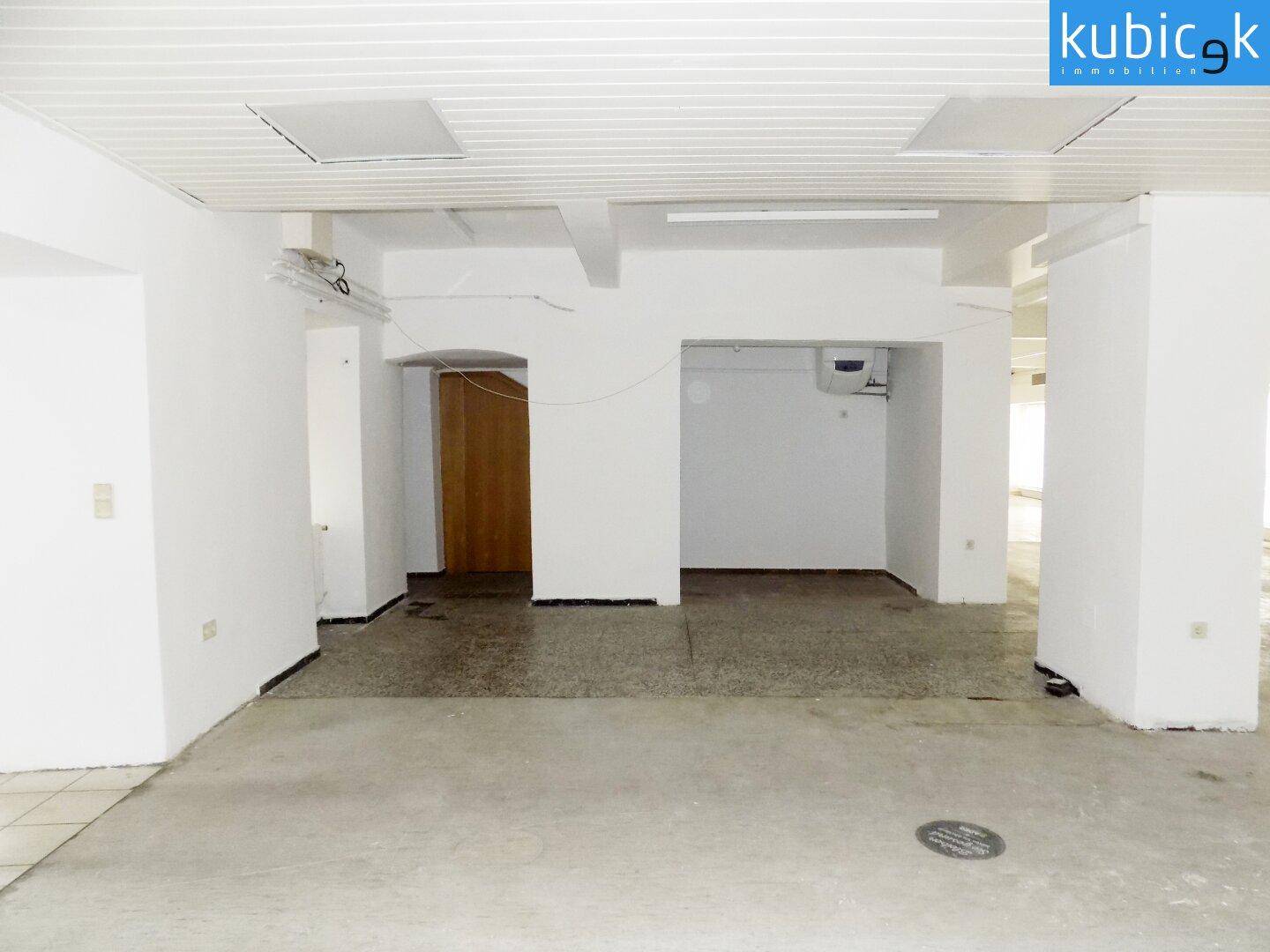 Gewerberaum mit gemischtem Bodenbelag aus Fliesen und Beton, weißen Wänden und einem Holztürrahmen.