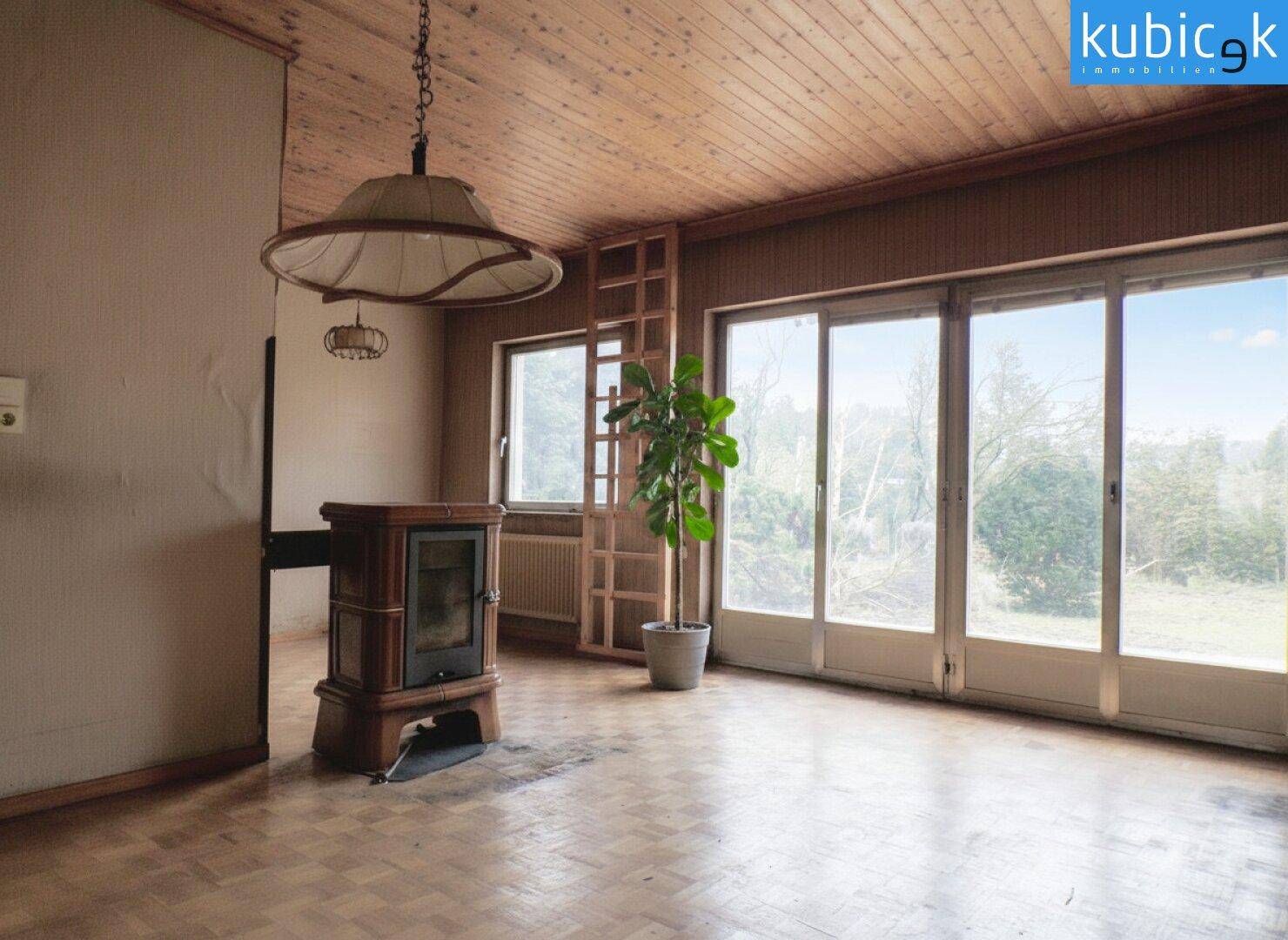Wohnzimmer mit Holzdecke, großen Fenstern und einem gemütlichen Kaminofen.