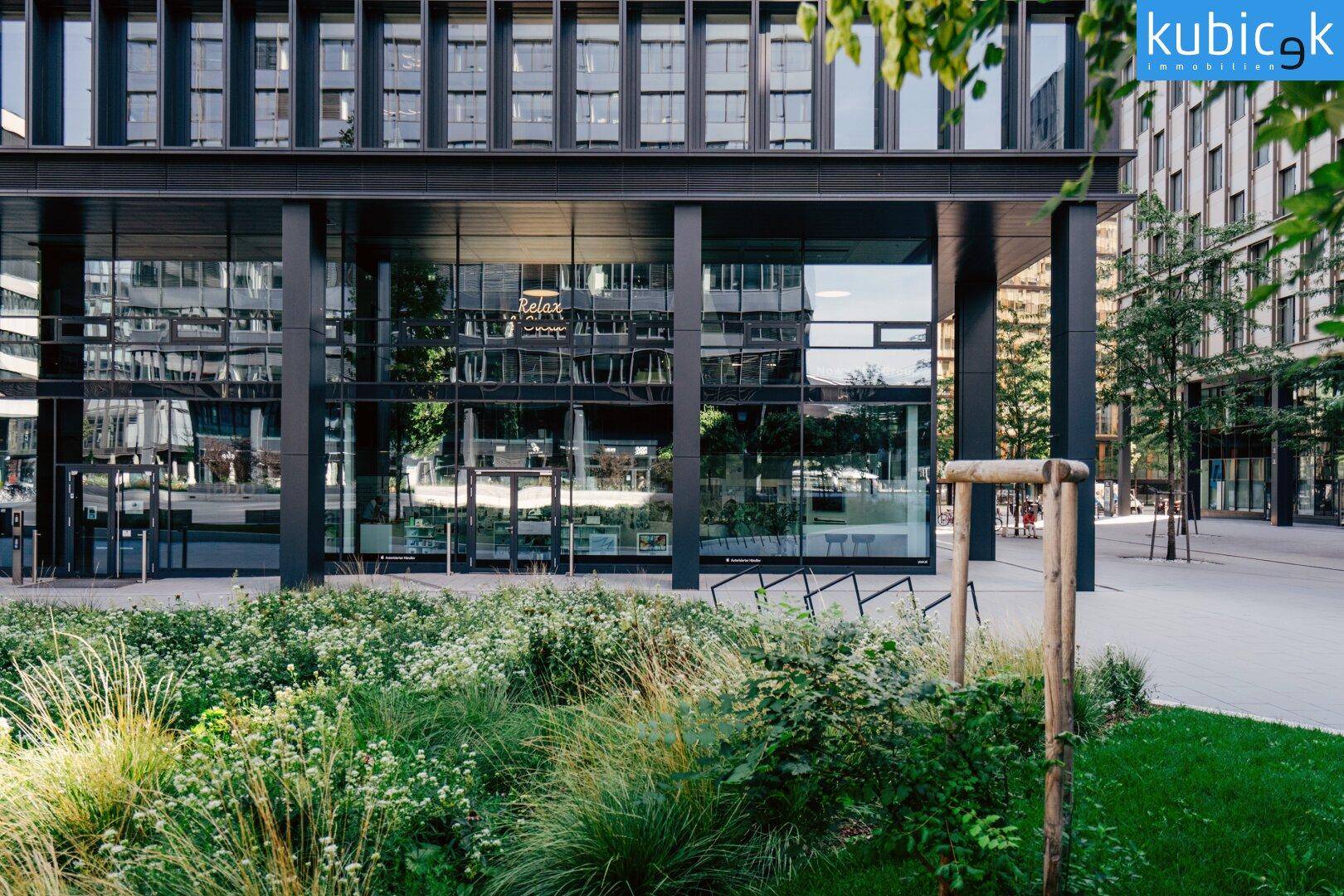 Eingangsbereich eines modernen Gebäudes mit üppiger Bepflanzung im Vordergrund und reflektierenden Fenstern.