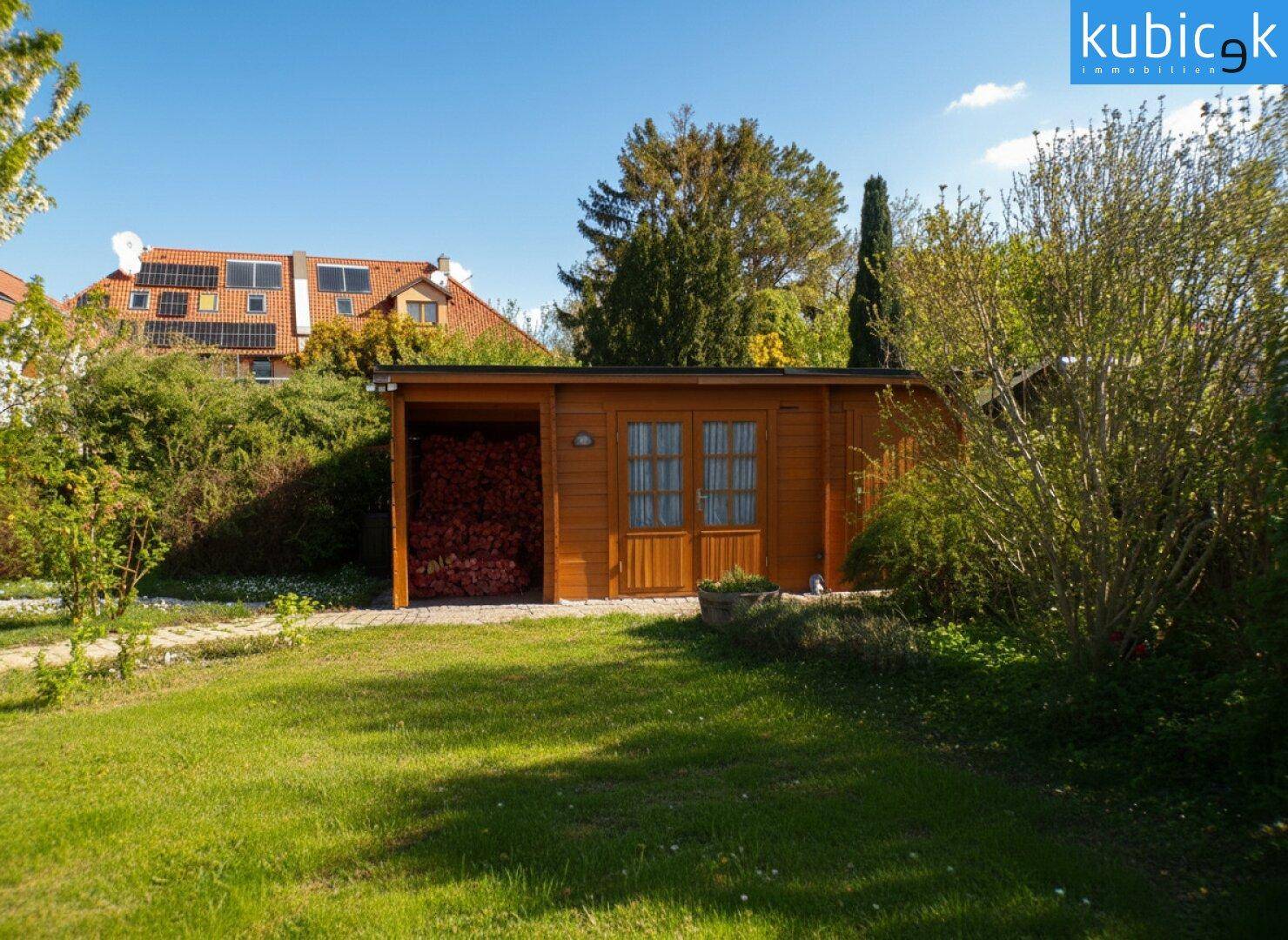 Holzgartenhaus mit überdachtem Lagerbereich für Brennholz im grünen Garten.