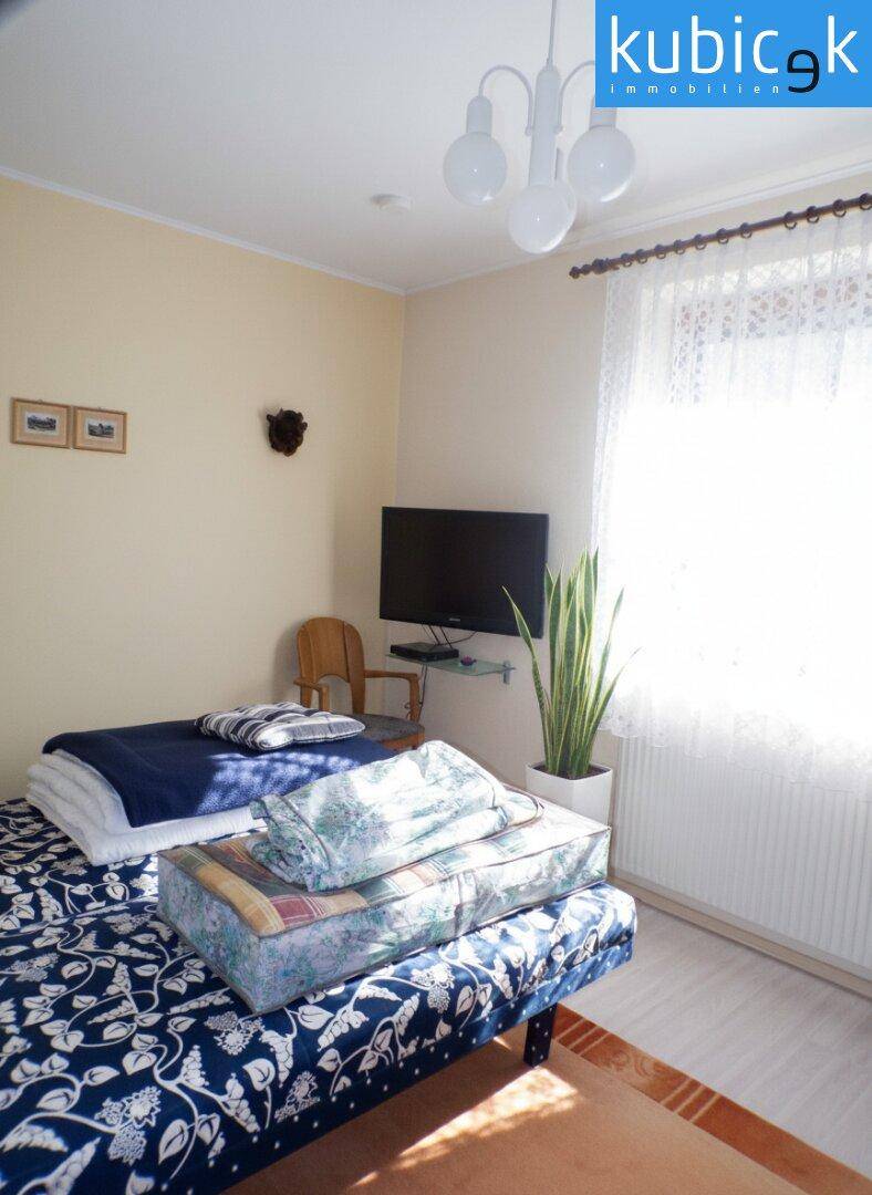 Helles Schlafzimmer mit Bett, Fernseher und großem Fenster für viel Tageslicht.