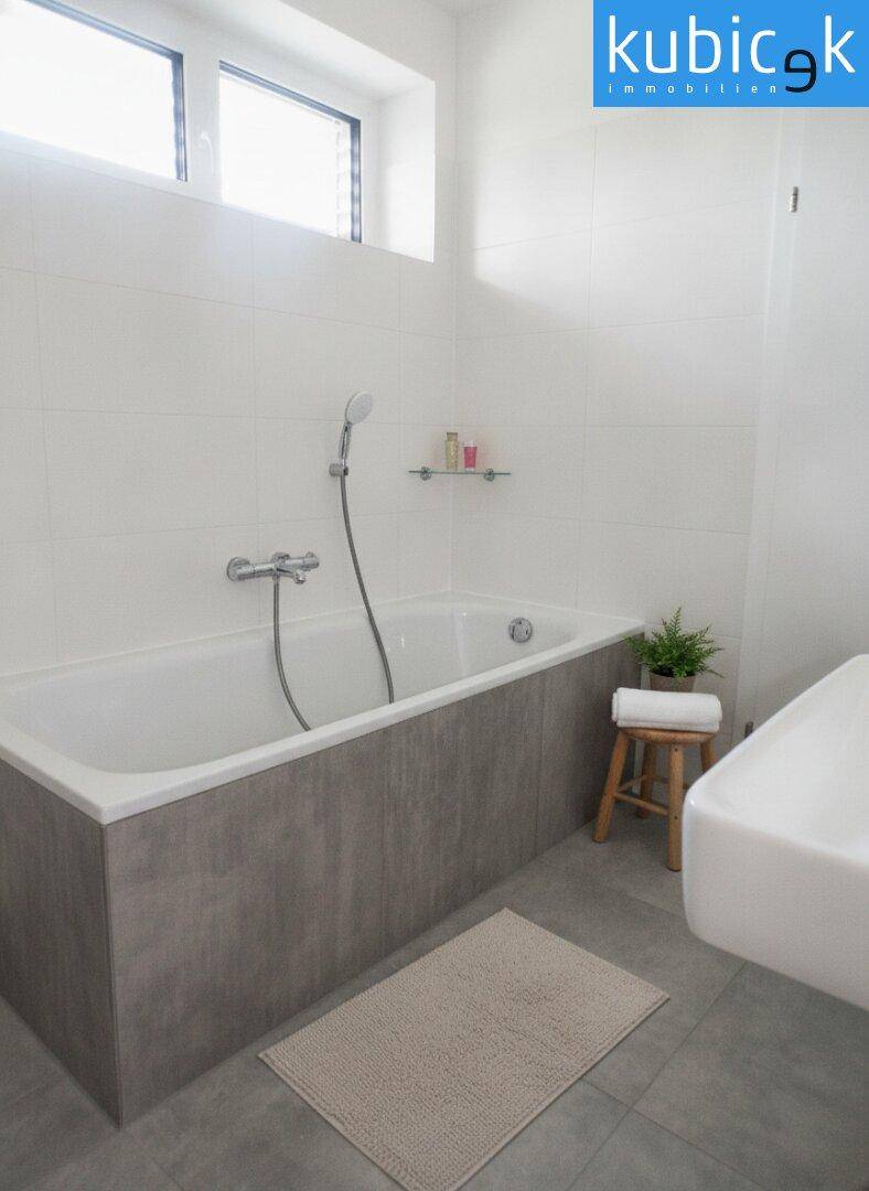 Zeitgemäßes Badezimmer mit Badewanne, Fenster und schlichten, hellen Fliesen.