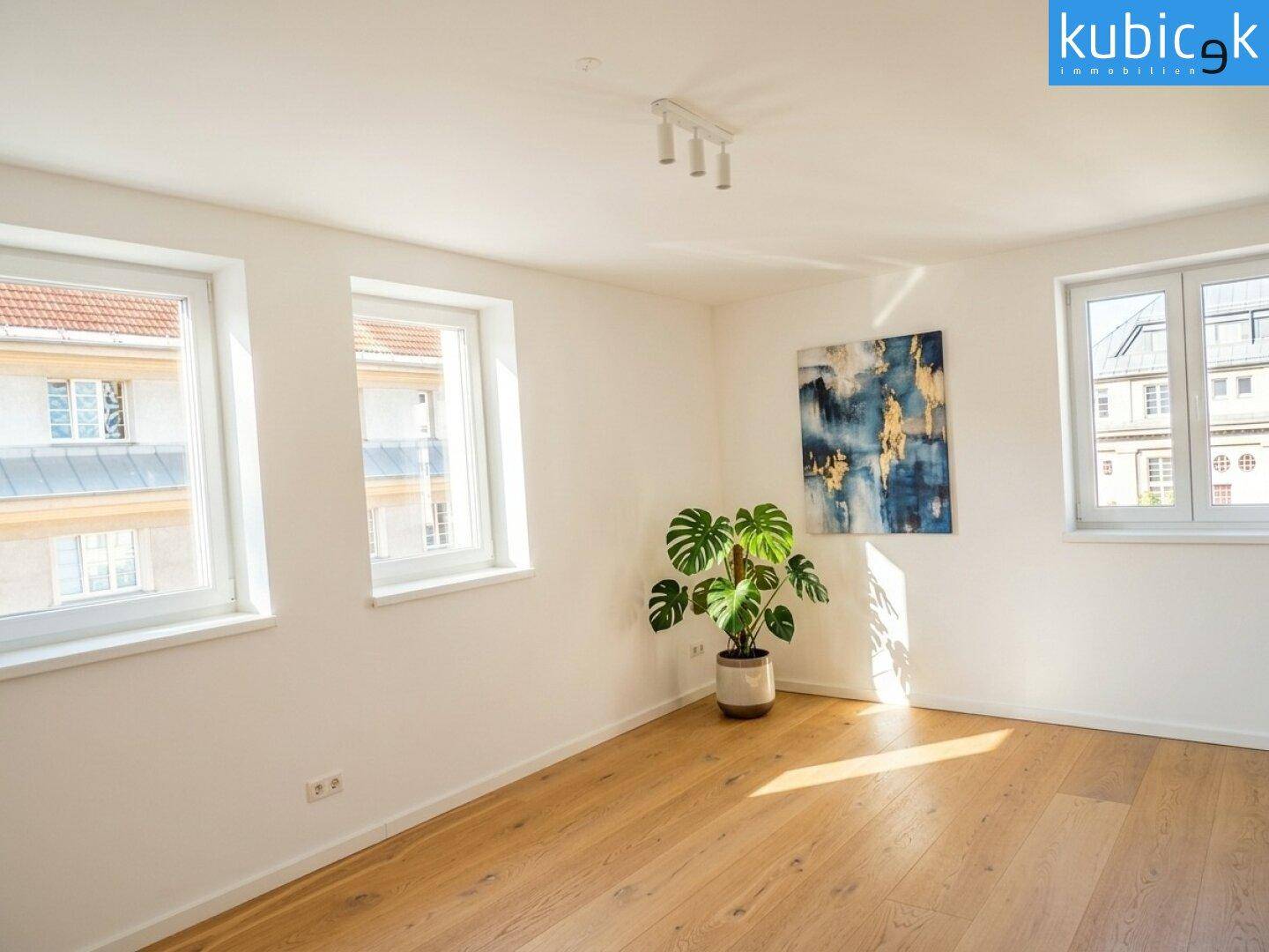 Sonniges Zimmer mit Holzboden und Blick auf die umliegenden Gebäude durch mehrere Fenster.