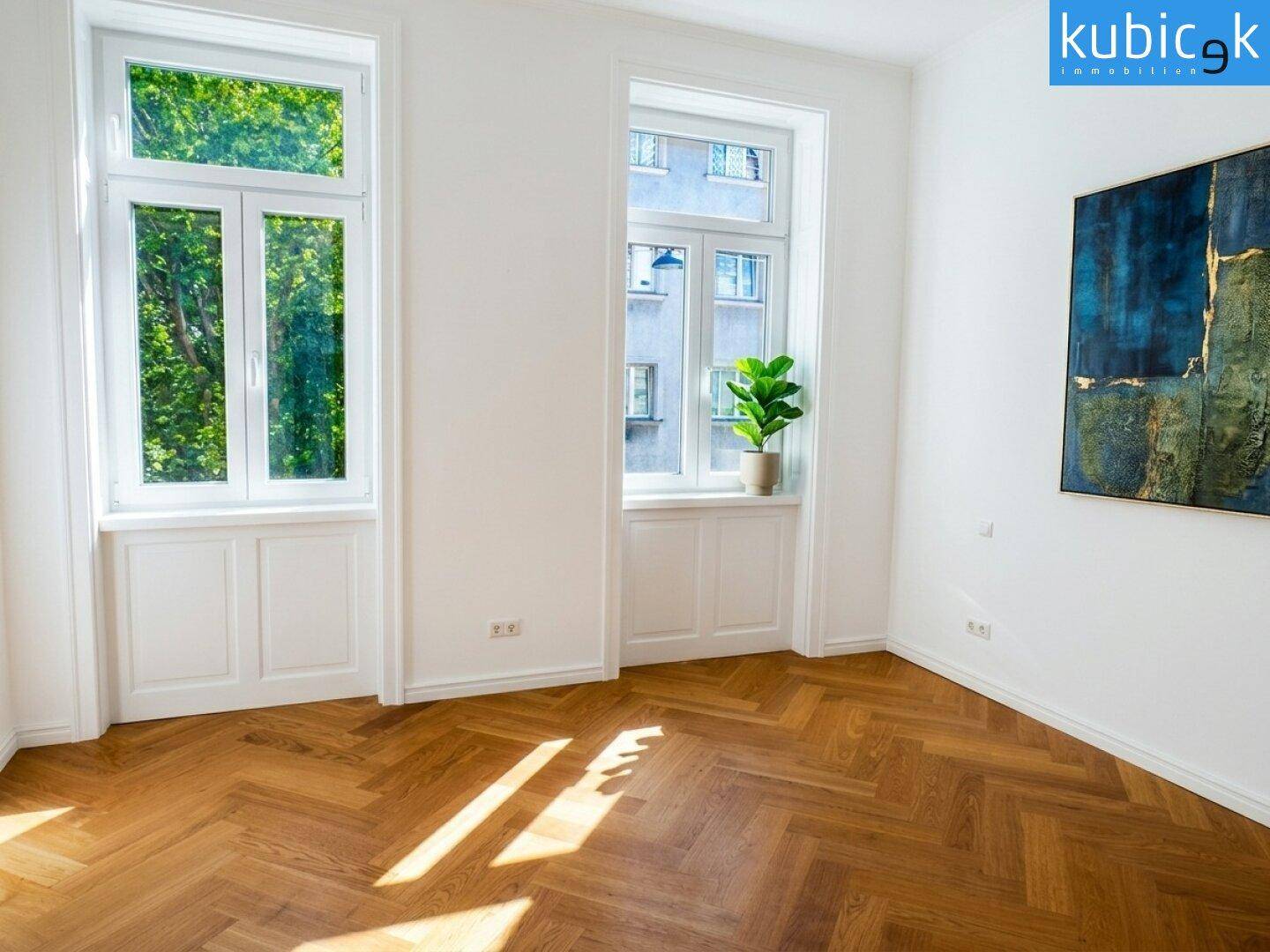Lichtdurchfluteter Raum mit elegantem Fischgrätparkett und großen Fenstern, ideal für Wohnzwecke.