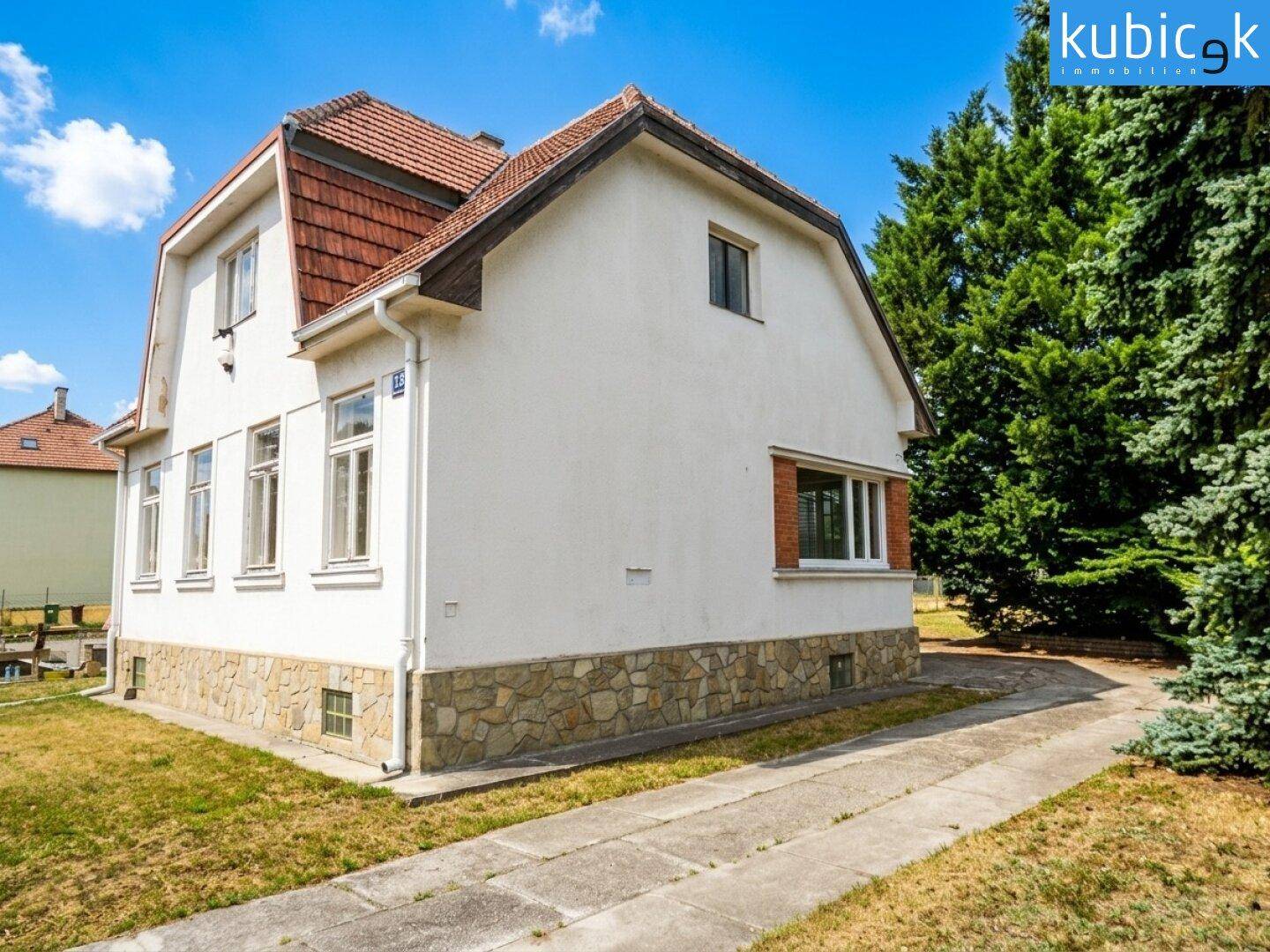 Ansicht eines Hauses mit weißer Fassade, Steinsockel und rotem Ziegeldach, umgeben von Grün.