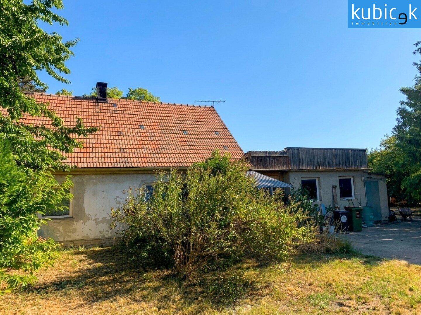 Ländliches Haus mit Ziegeldach, umgeben von Bäumen und Grünflächen unter blauem Himmel.
