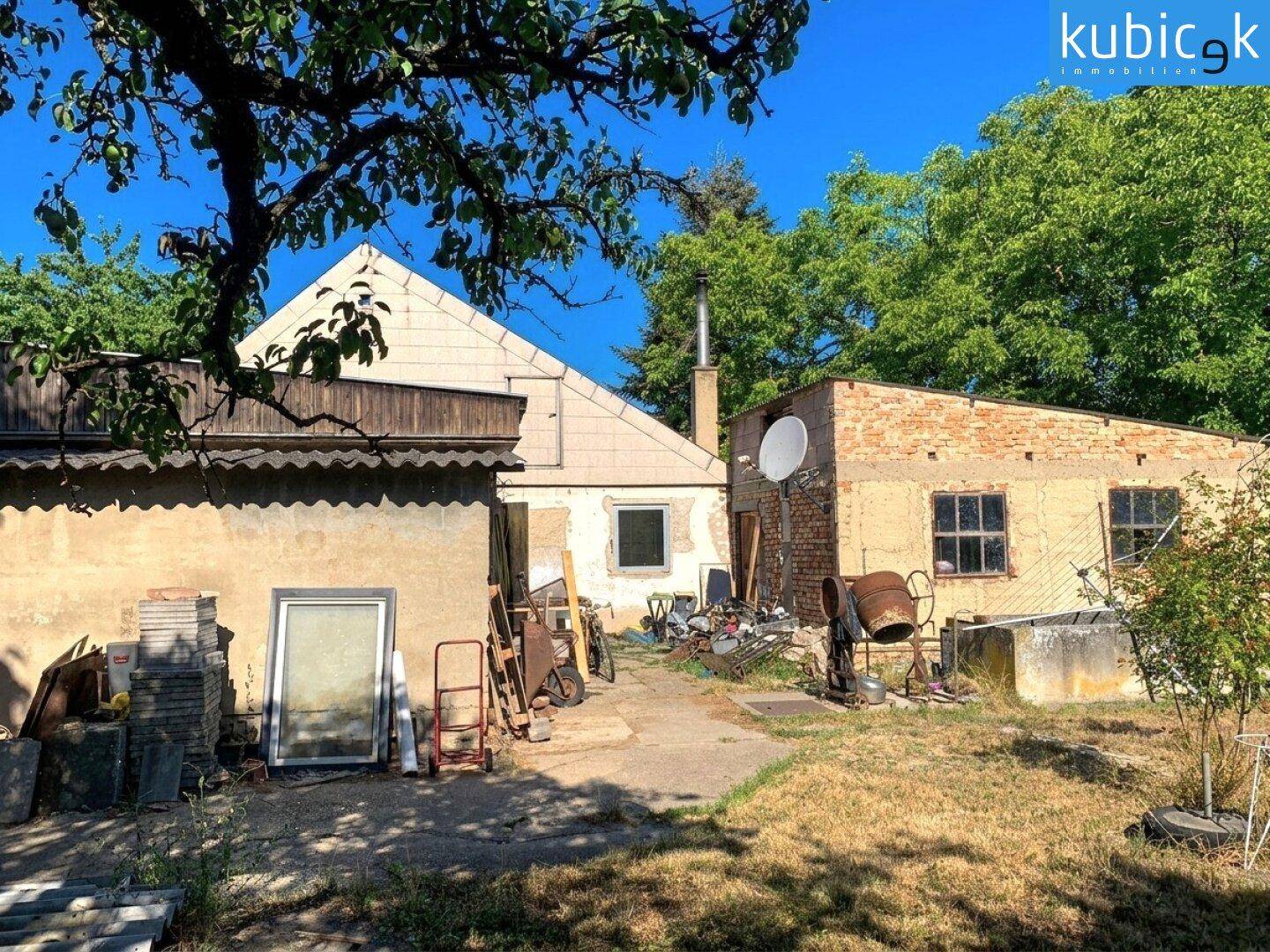 Rückseite eines älteren Hauses mit Nebengebäuden und Baumaterialien im ungepflegten Gartenbereich.