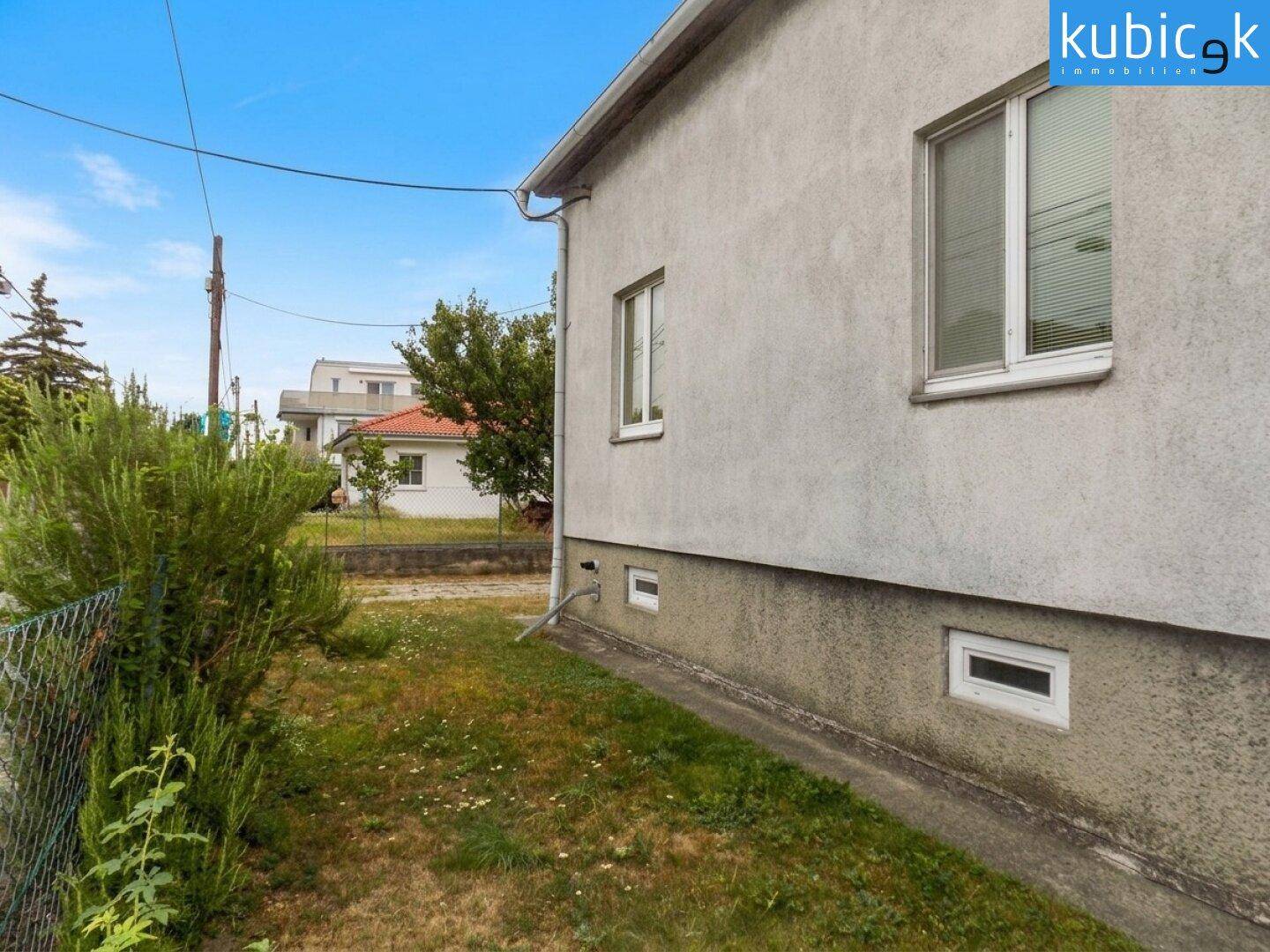 Seitenansicht des Hauses mit kleinen Kellerfenstern und einer Hecke im Vordergrund.